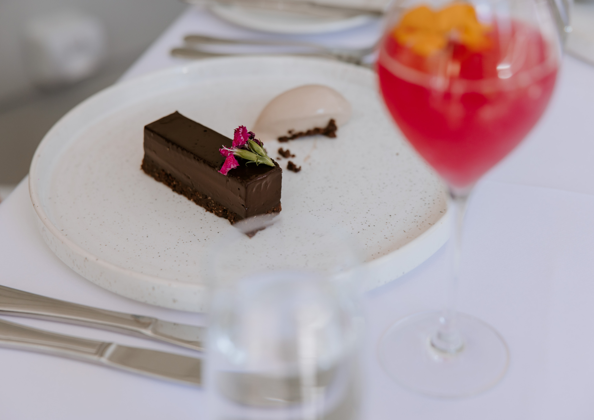A slice of chocolate cake with a pink flower on top, a scoop of ice cream, and a glass of red fruit cocktail on a white table.