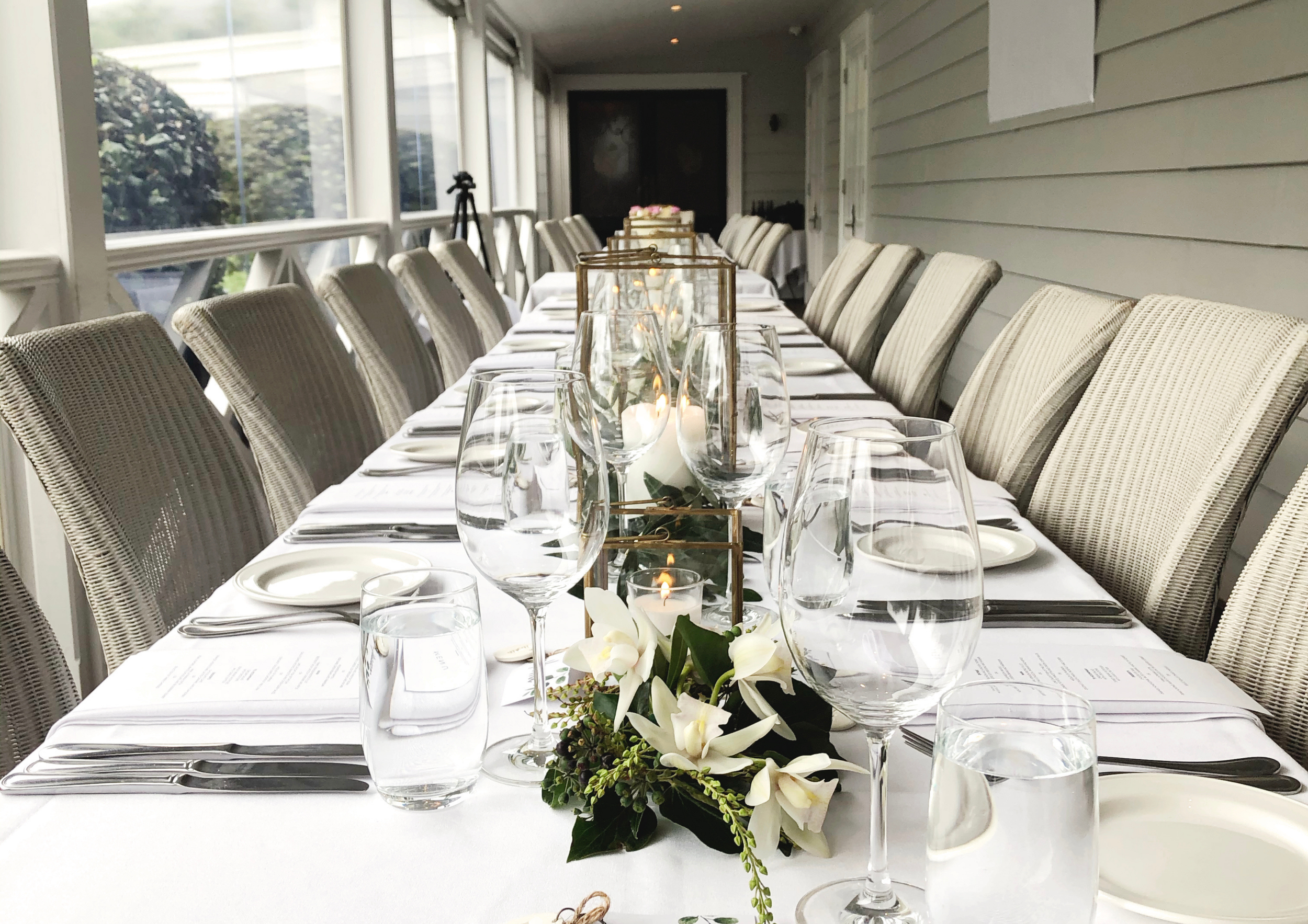 Elegant dining table arranged for a formal event with white tablecloth, wine glasses, floral centerpiece, candles, and silverware in a sunlit room.