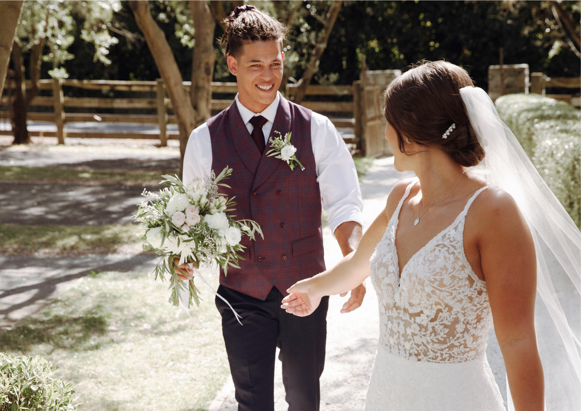 A groom in a plaid vest and tie holding a bouquet of flowers, smiling at a bride in a lace wedding dress with a veil, outdoors in a garden setting.