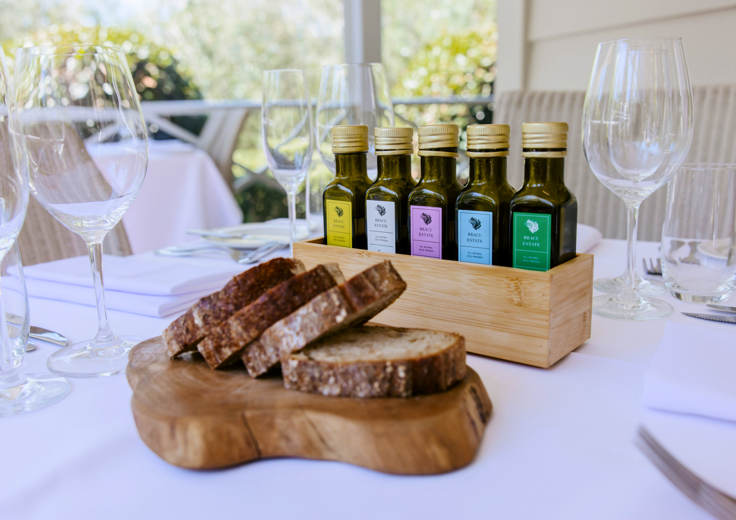 A dining table set outdoors featuring a wooden board with sliced bread, a wooden box containing several small bottles of flavored oil or vinegar, and empty wine glasses.