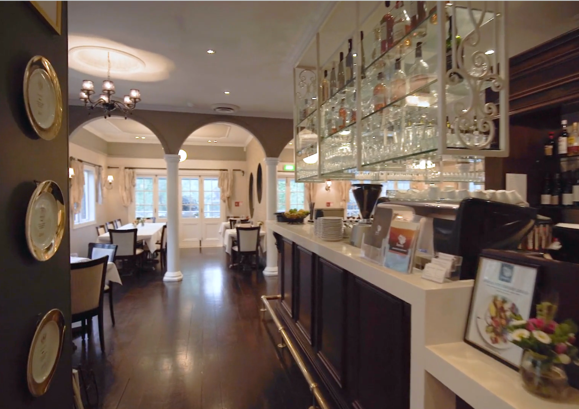 Elegant dining area with white tablecloths, dark wooden chairs, and large windows with curtains. A bar area with glassware hanging overhead and various bottles displayed. Soft lighting and chandelier illuminate the room.