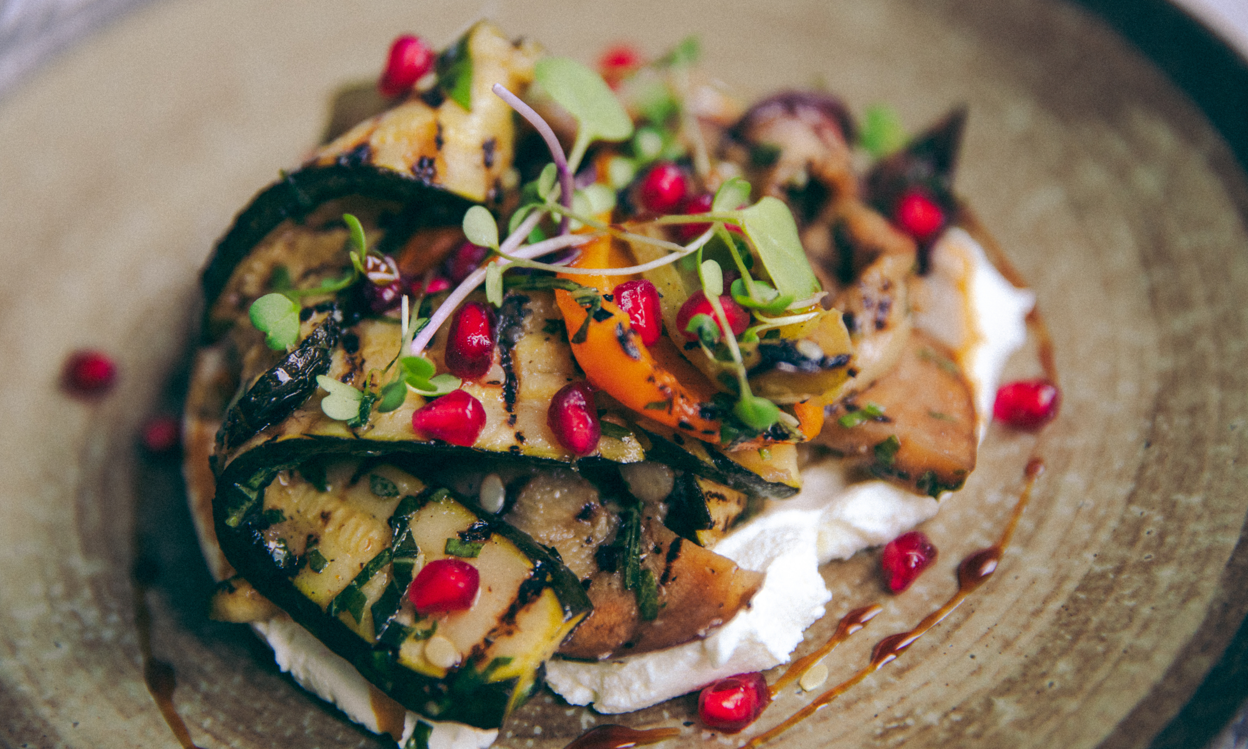 Grilled vegetable dish with pomegranate seeds, microgreens, and sauce on a textured brown plate.