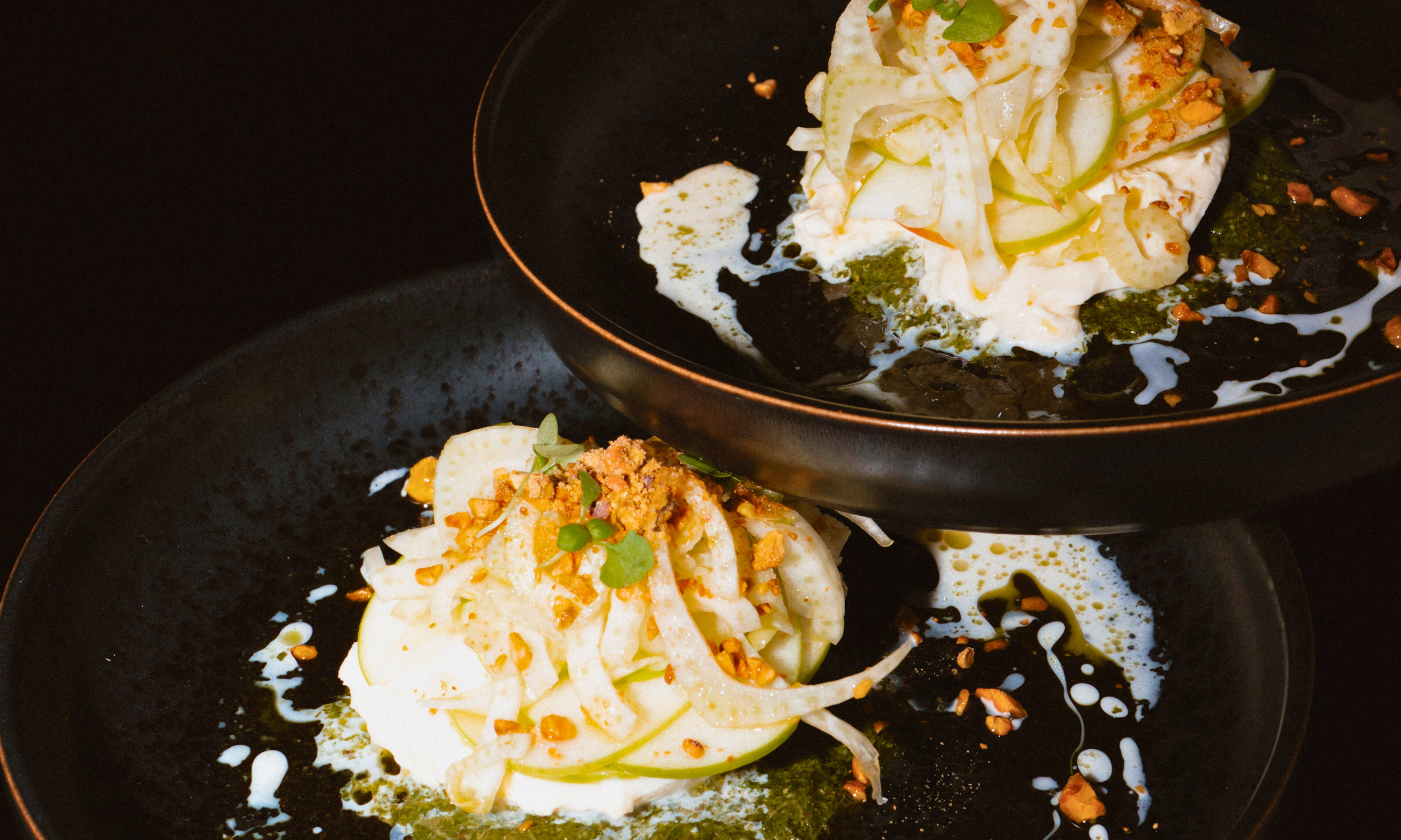Two black plates with zucchini noodles topped with crushed nuts, herbs, and a sauce, with some sauce and nuts spilled on the plates.