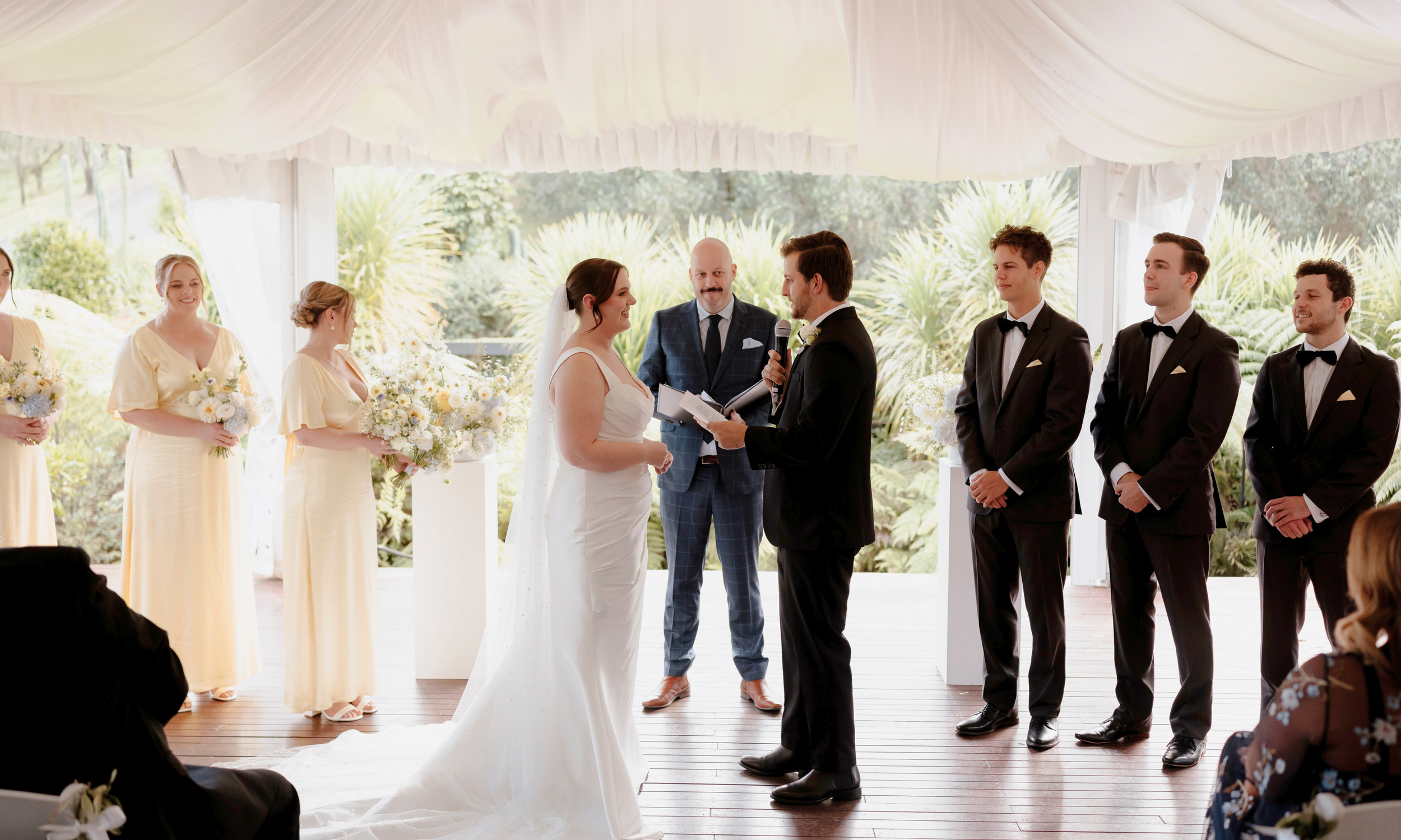 A wedding ceremony with a bride and groom exchanging vows inside a decorated venue with large windows and lush greenery outside.