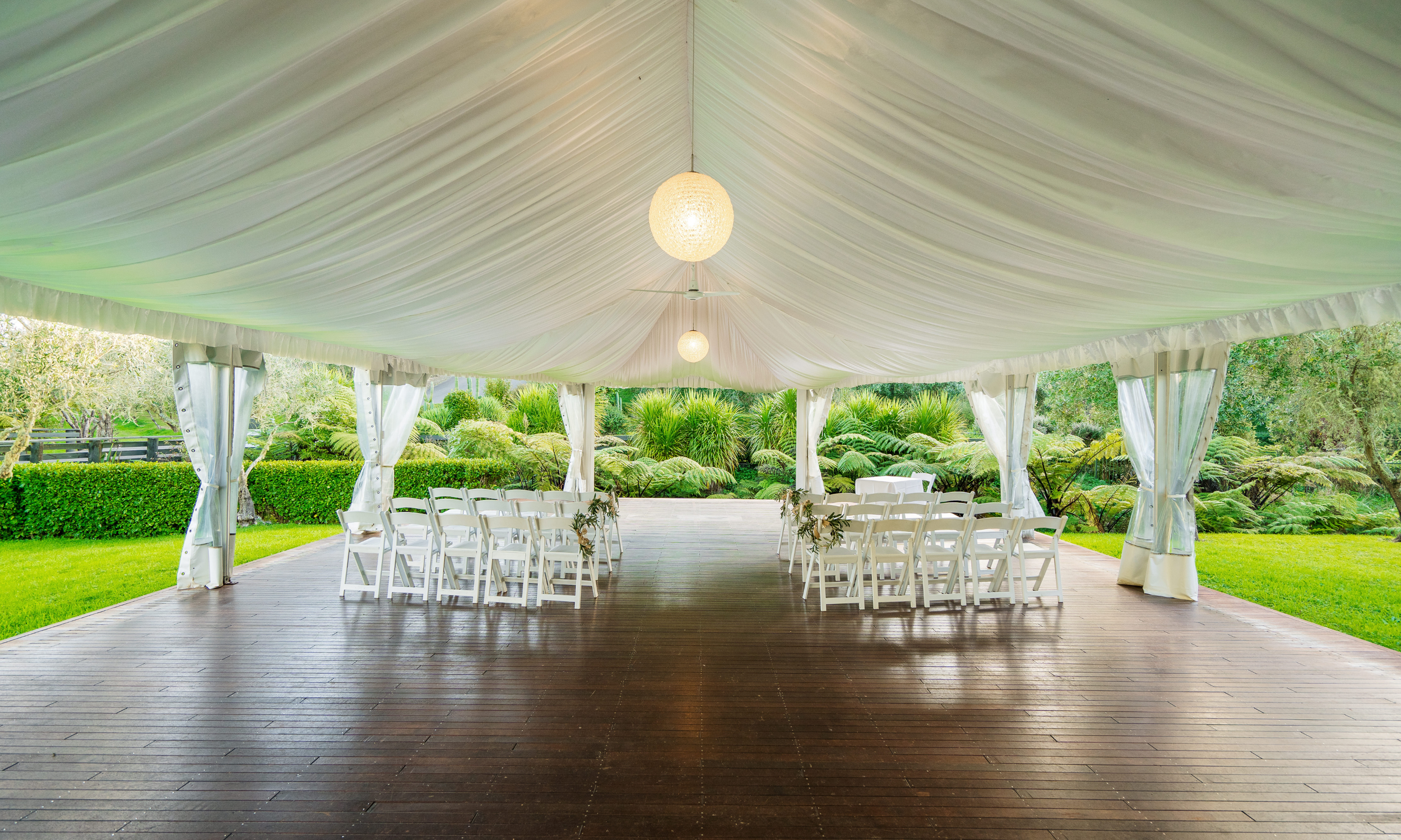 Wedding ceremony setup under a large white tent with white chairs arranged in rows, decorated with small floral arrangements, on a wooden deck with a green garden background.