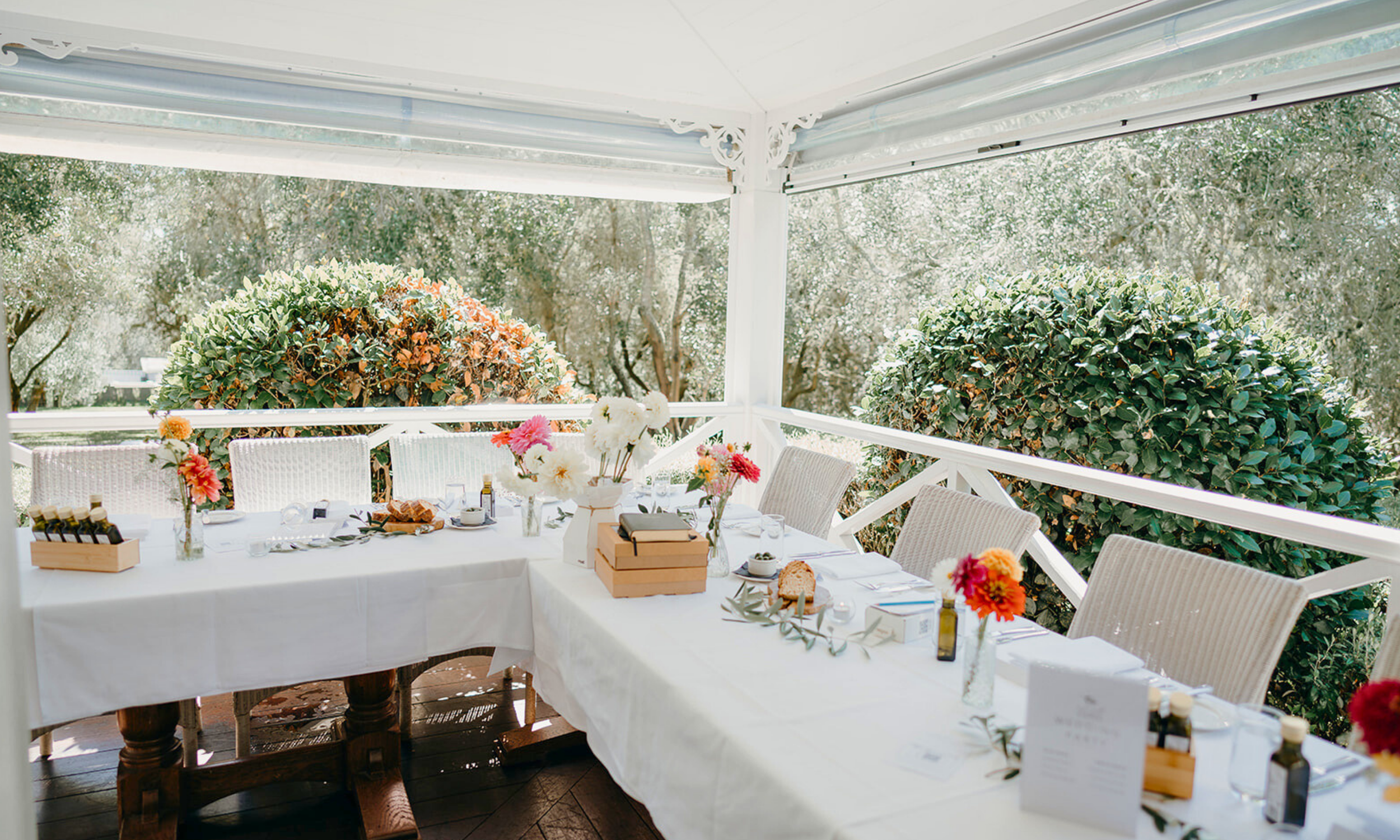 A decorated outdoor dining table with white tablecloth, floral centerpieces, wine bottles, and chairs, surrounded by lush green bushes and trees.