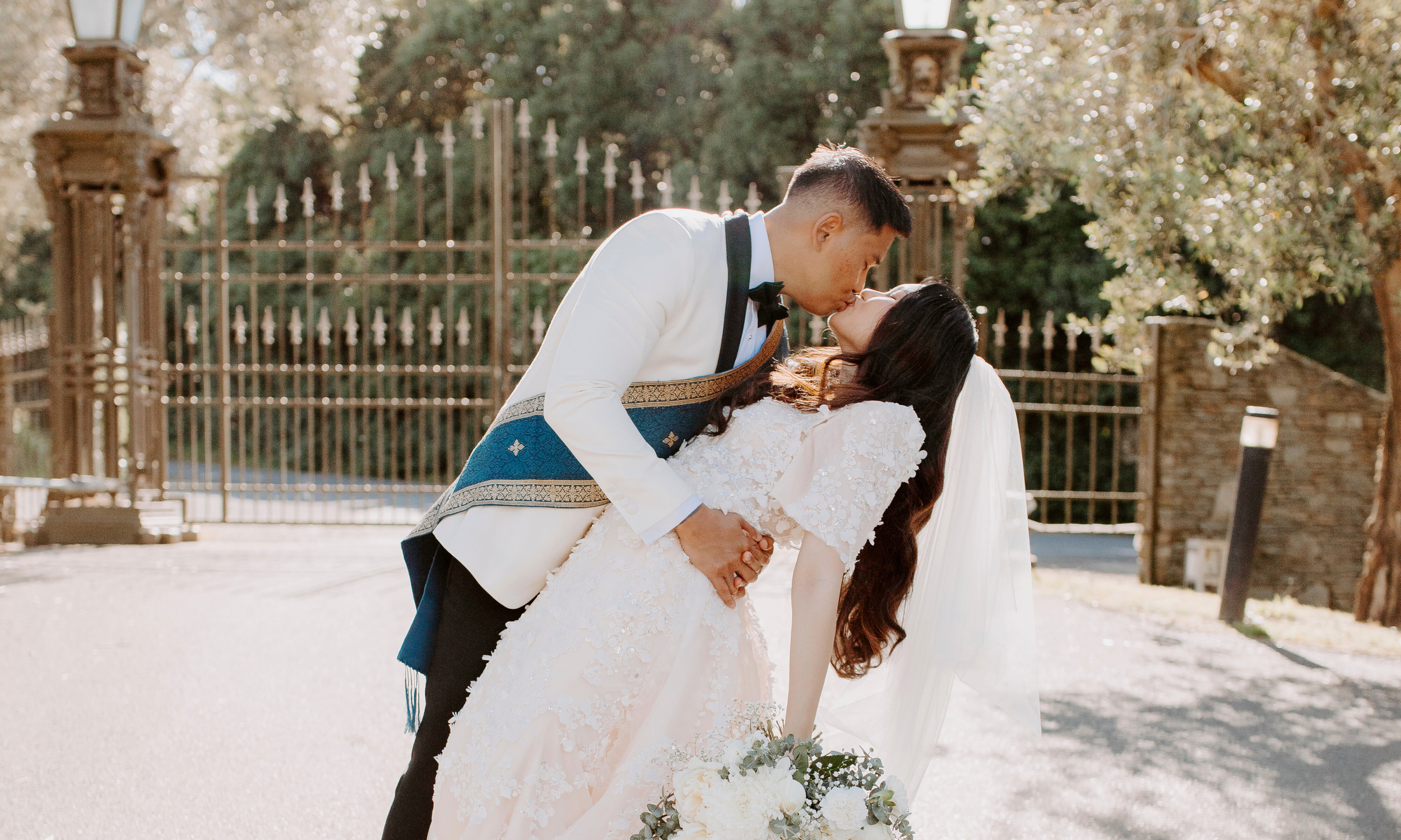 A newlywed couple sharing a kiss outdoors during daytime, with the groom dipping the bride. The bride is holding a bouquet of white flowers, and the scene includes a decorative gate and trees in the background.