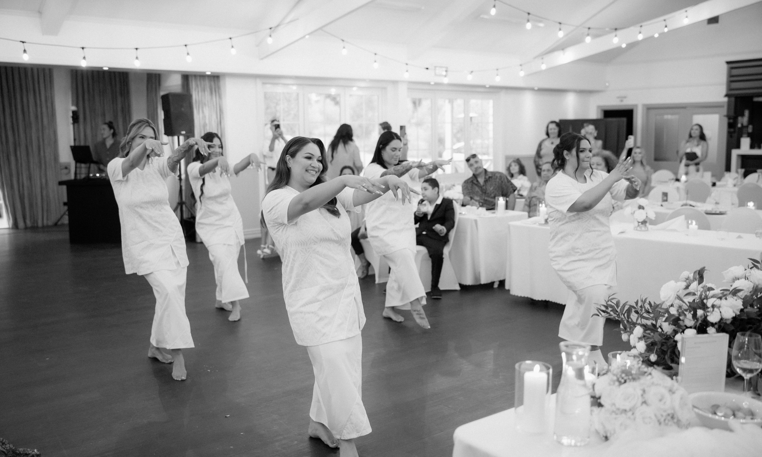 A group of women in white dresses perform a dance at a celebration, with guests seated at tables watching and taking photos in a decorated hall.