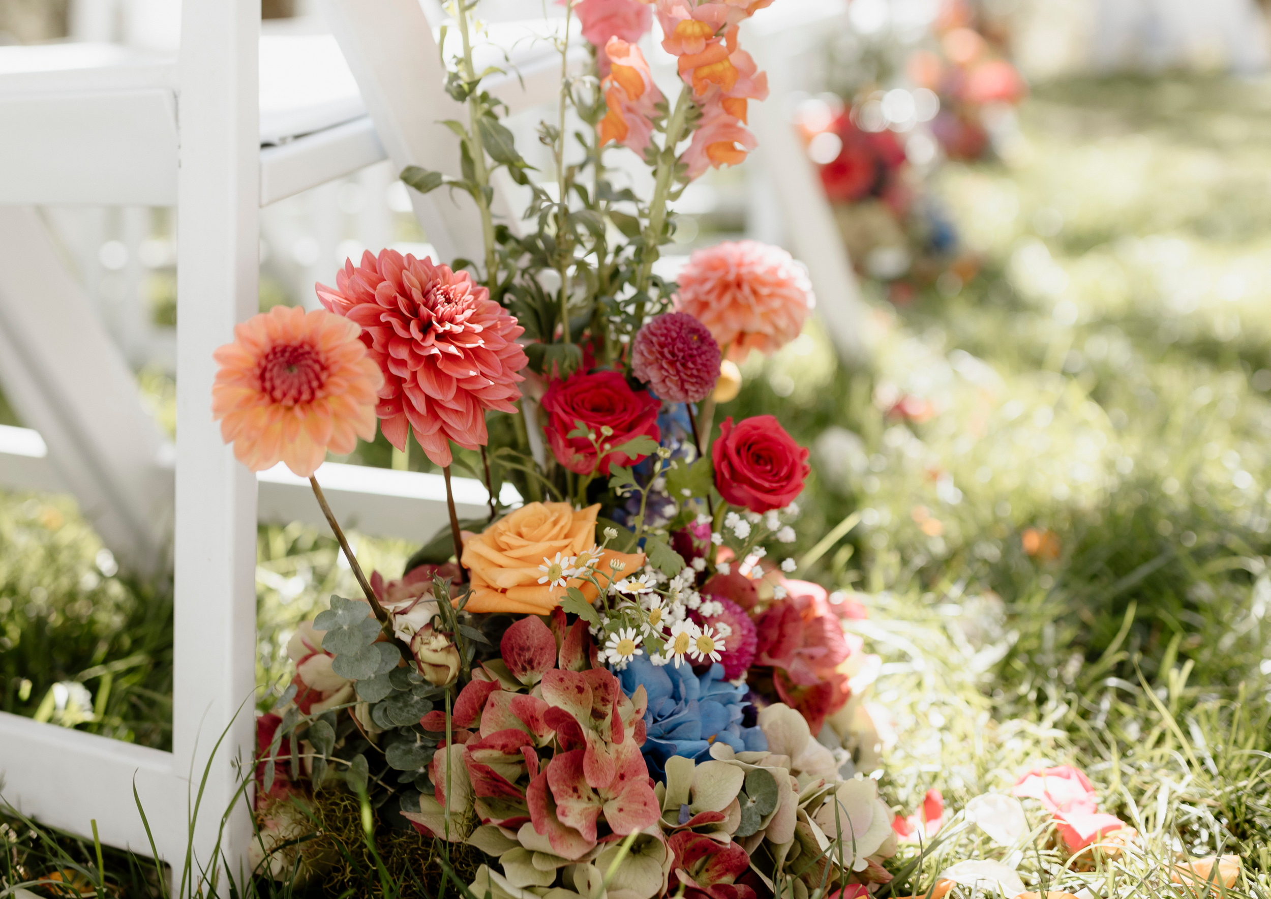 Colorful bouquet of flowers including roses, daisies, hydrangeas, and dahlias on grass near a white wooden chair with blurred flowers in the background.