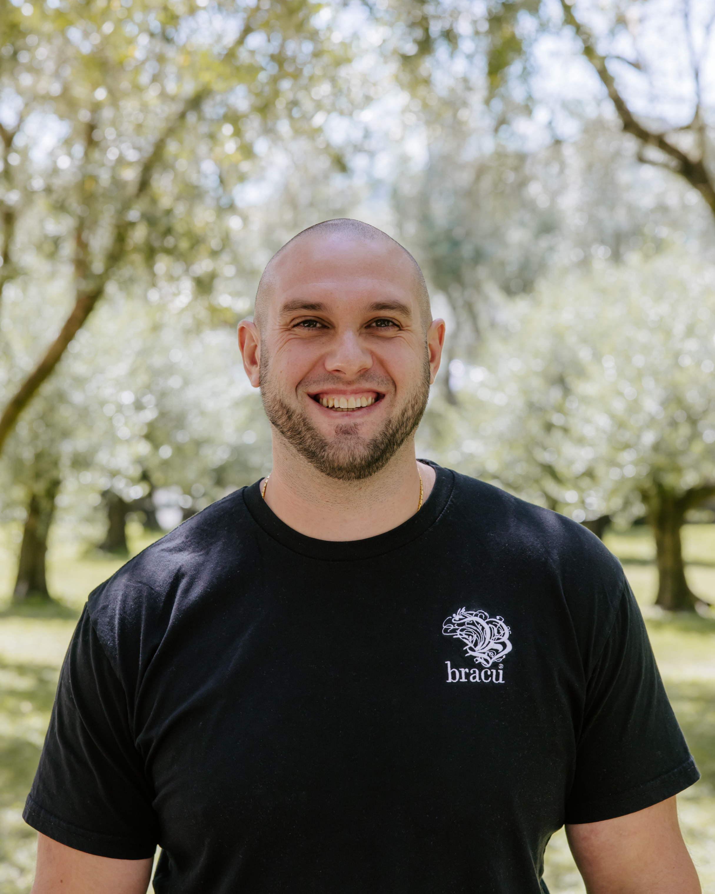 A smiling man with a beard and short hair wearing a black t-shirt with a white logo, standing outdoors in a park with trees in the background.