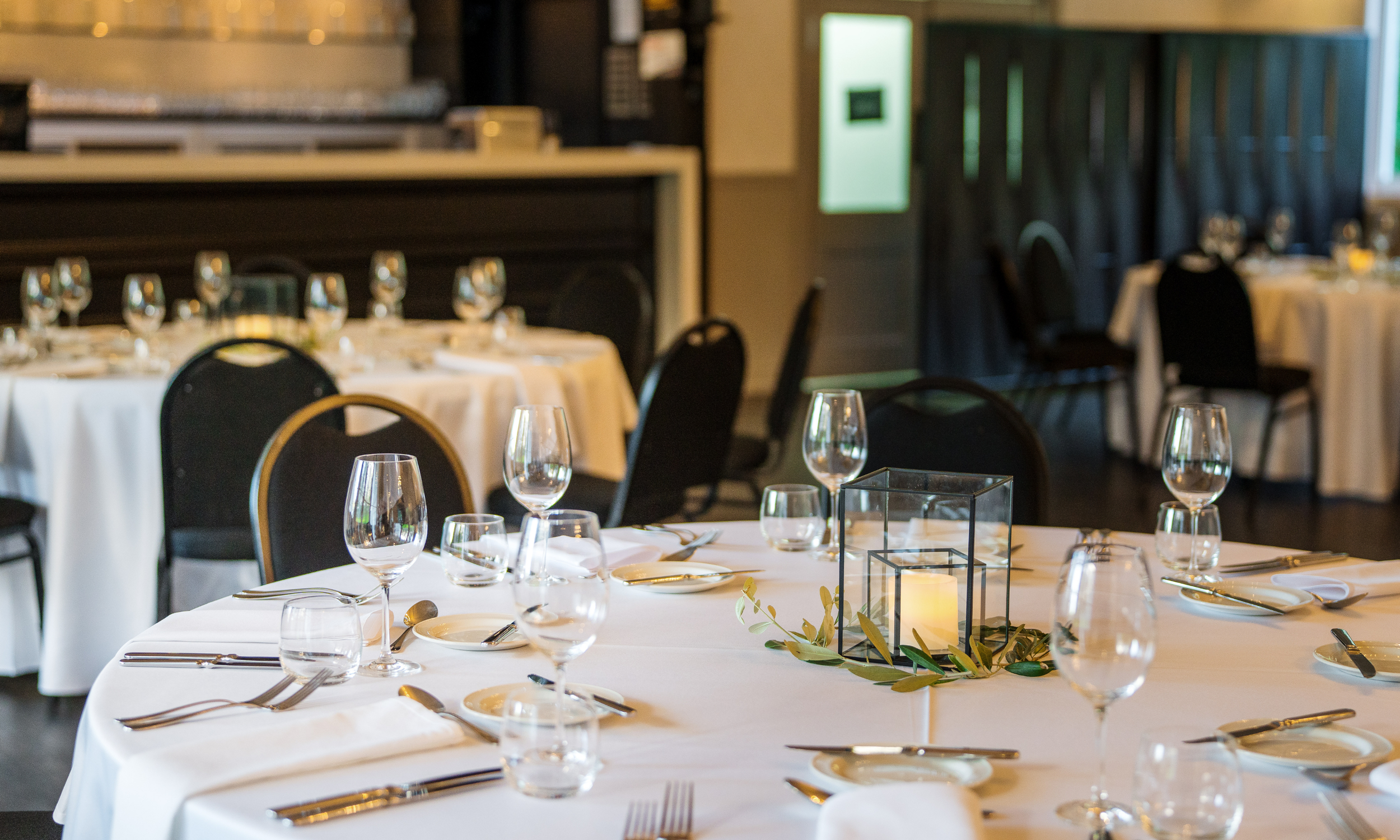 A round dining table set for a formal event with white tablecloths, wine glasses, silverware, and a decorative centerpiece with a candle inside a glass box, surrounded by green leaves.