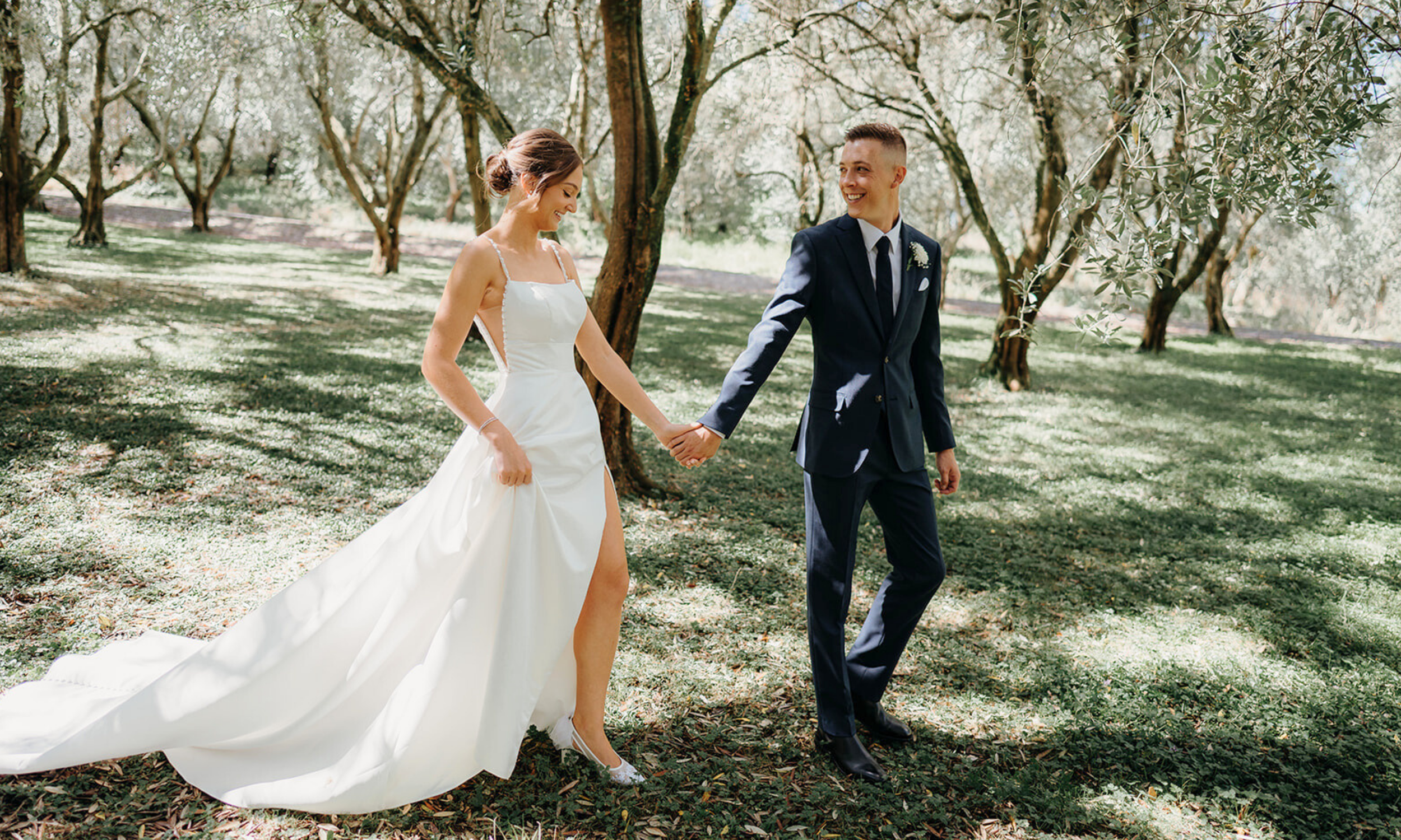 A newlywed couple holding hands and walking through a lush, green orchard with trees casting dappled sunlight. The bride is wearing a white wedding gown with a high slit, and the groom is in a dark suit.