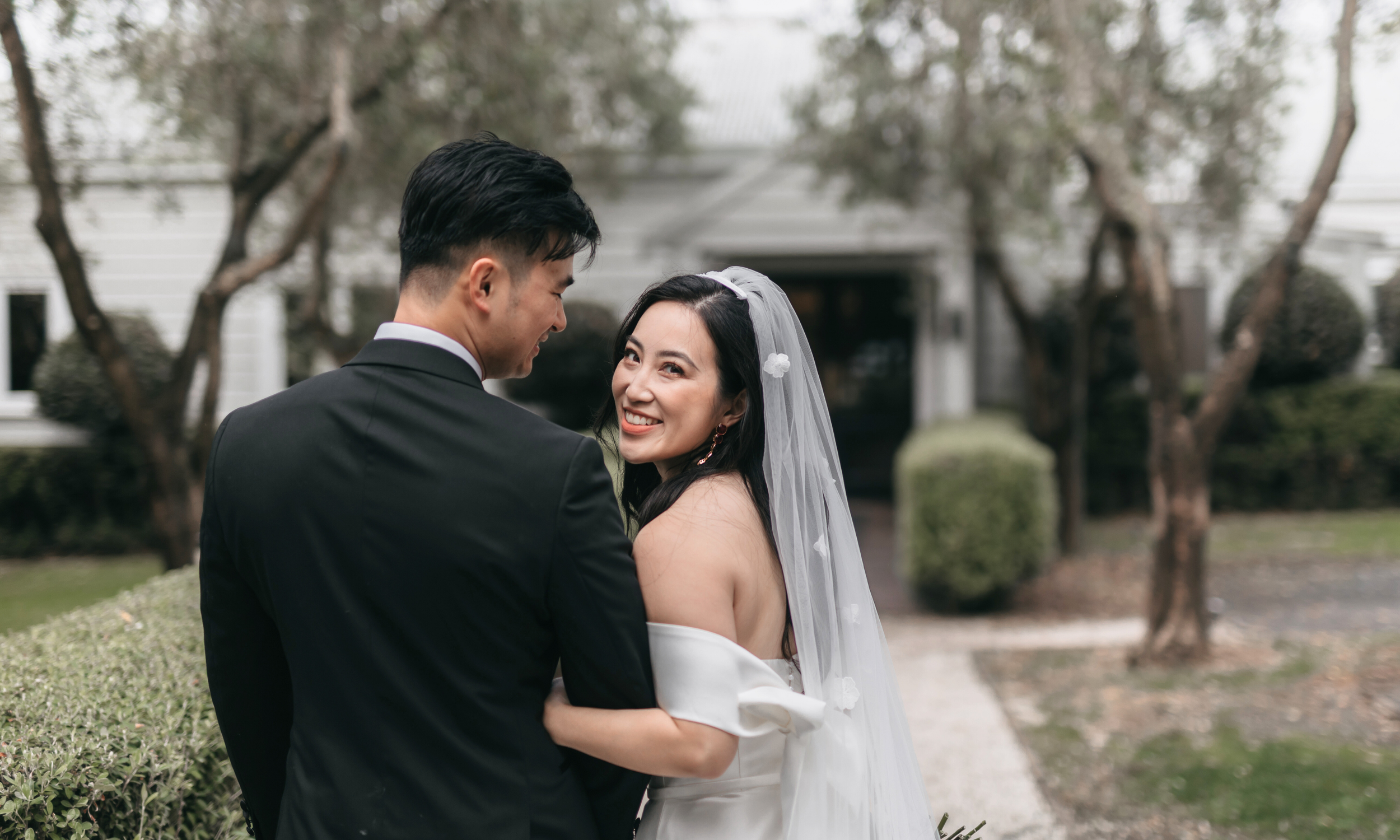 A bride and groom standing outside on a cloudy day, smiling at each other. The bride is wearing a white off-the-shoulder wedding gown with a veil, and the groom is dressed in a black suit. There are trees and bushes in the background.