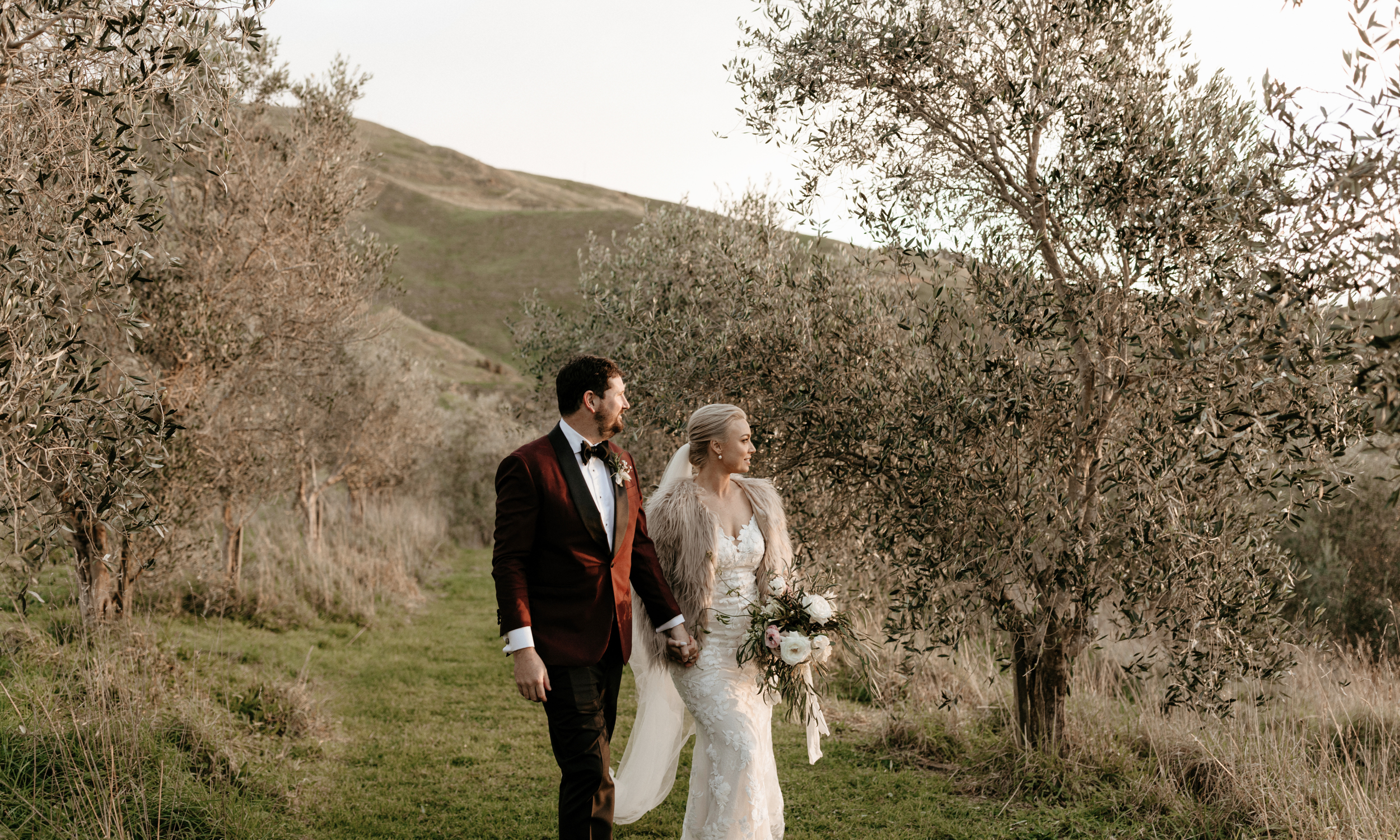 A bride wearing a white wedding dress and fur jacket holding a bouquet of flowers, and a groom in a dark red velvet tuxedo with a black bow tie, walking hand in hand outdoors through a grassy area with trees and hills in the background.
