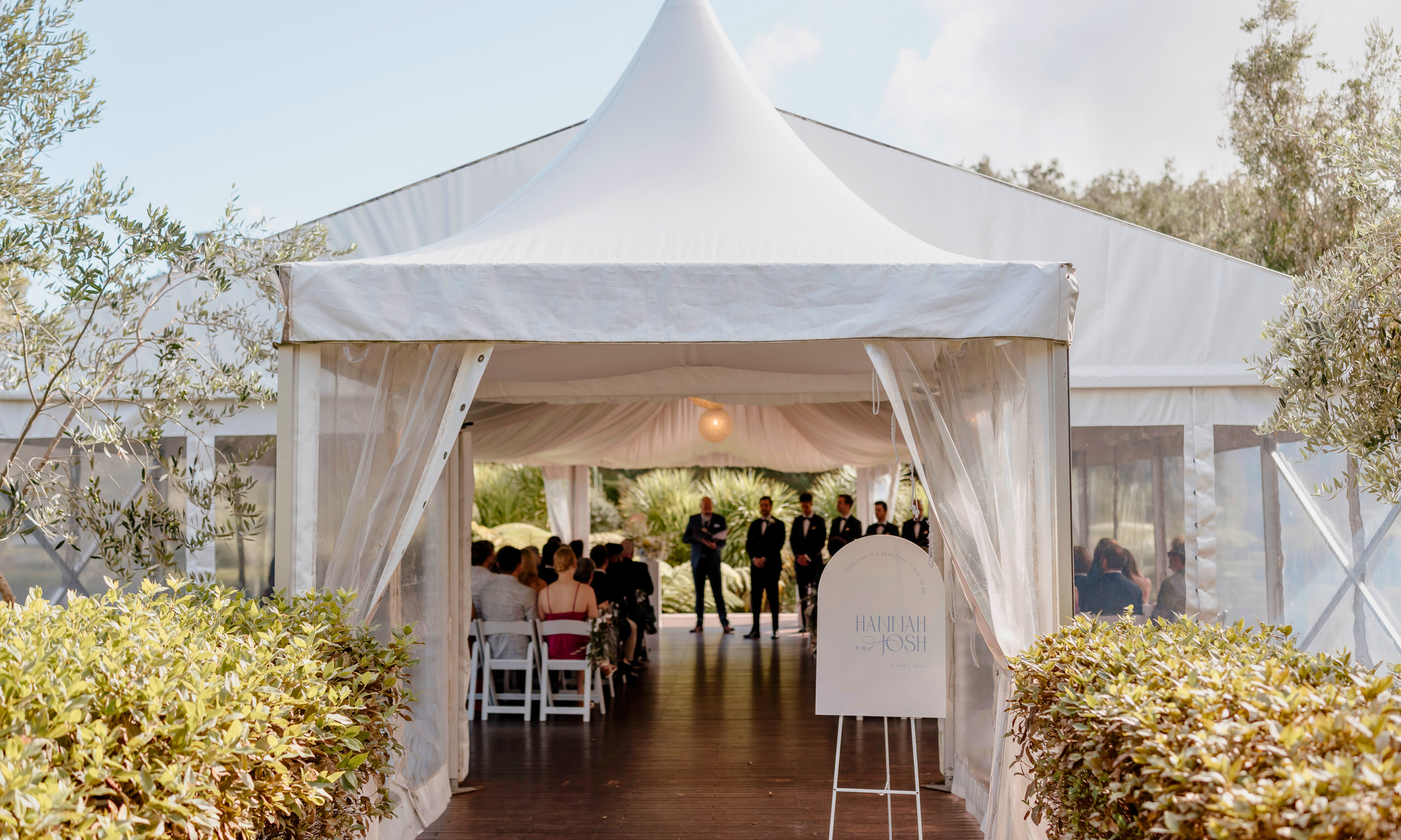 Outdoor wedding ceremony in a white tent with guests seated, and a group of groomsmen standing at the front, with a sign reading 'Hannah and Josh'.