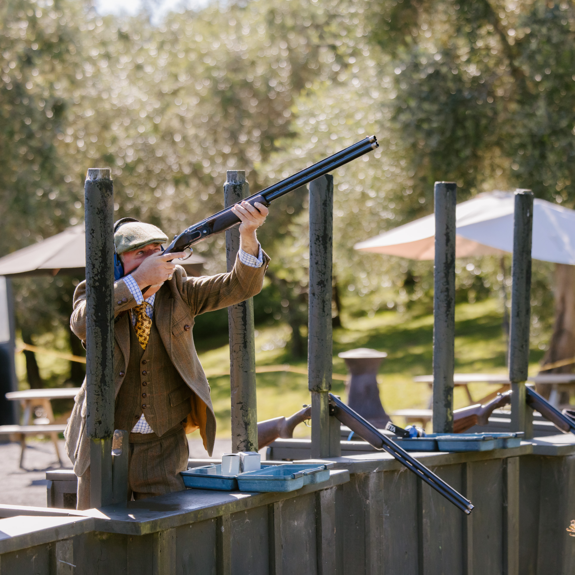 A man in vintage clothing shooting a double-barrel shotgun at a clay target range outdoors at Bracu Estate, with trees and umbrellas in the background.