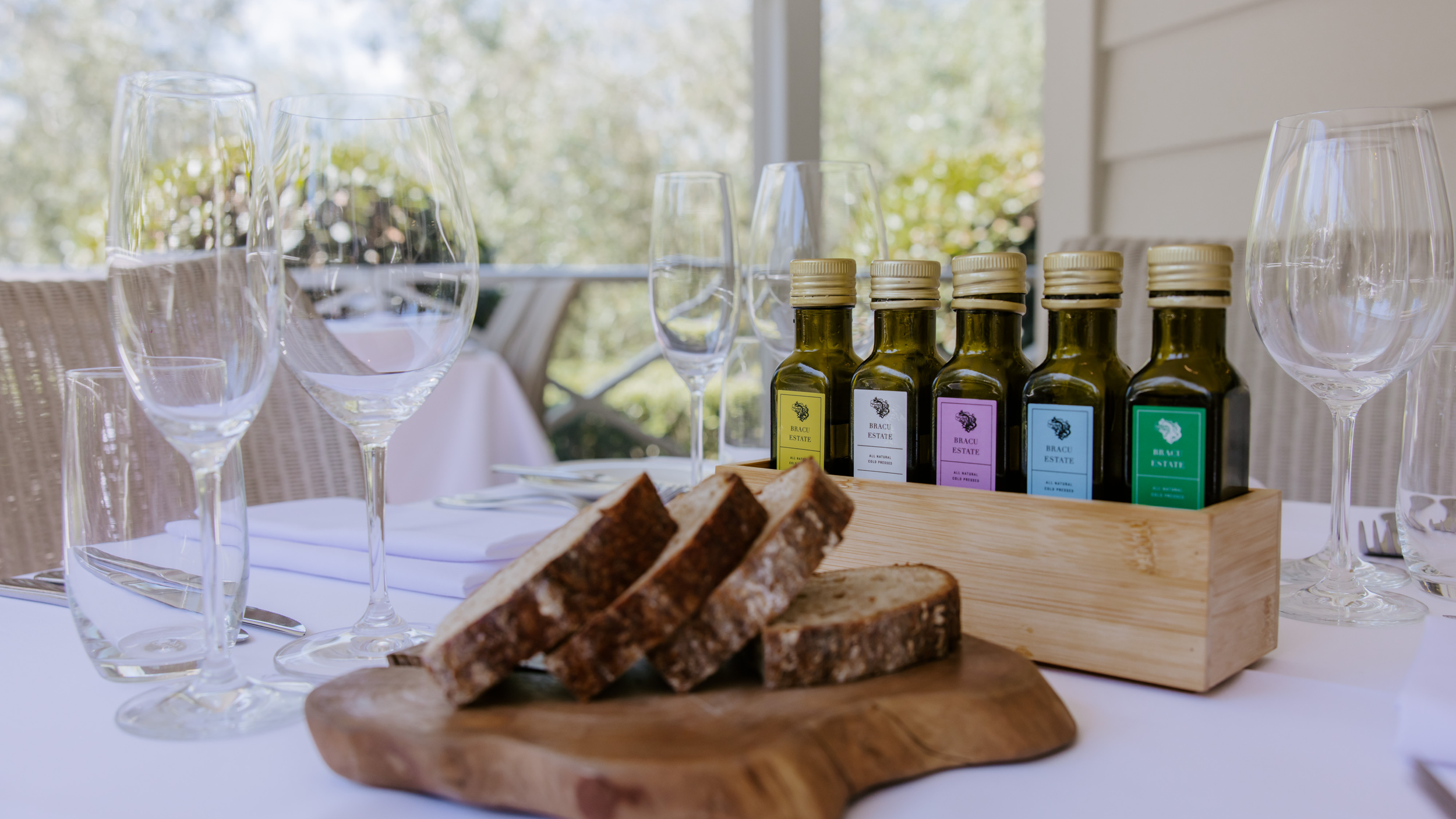 Table set for a wine tasting with empty wine glasses, slices of bread on a wooden board, and bottles of wine labeled 'Bracu Estate' in a wooden box.