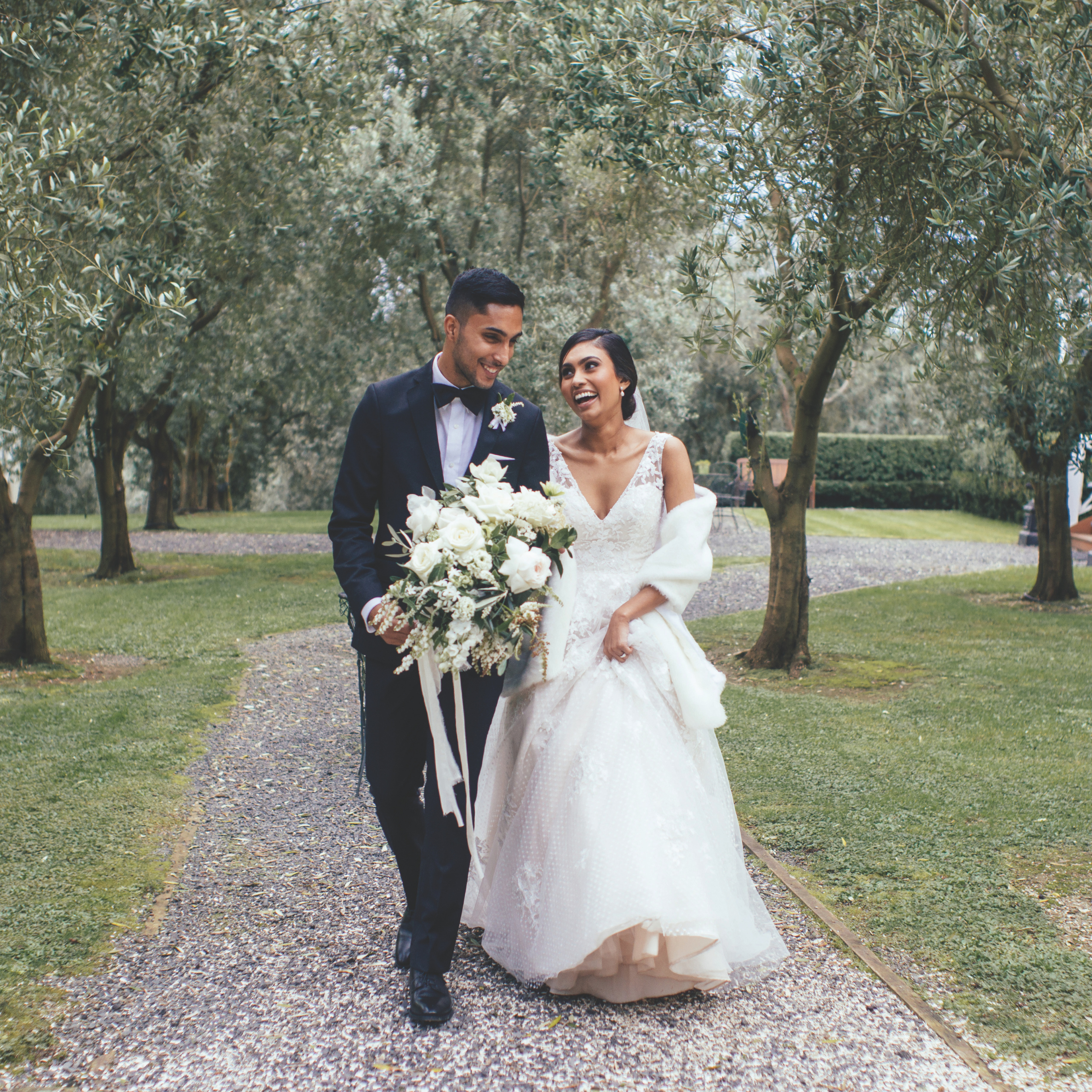 A happy bride and groom walking outdoors at Bracu Estate surrounded by olive trees. The bride is wearing a white wedding gown and holding a bouquet of white flowers, while the groom is dressed in a dark suit with a white shirt and bow tie.