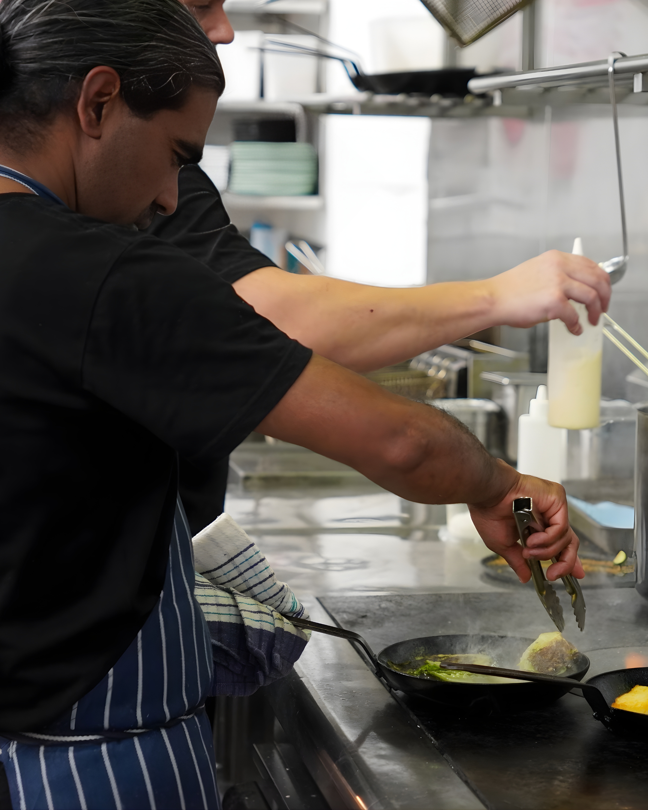 Two chefs cook on a stovetop in a professional kitchen, one using tongs to handle food in a frying pan.
