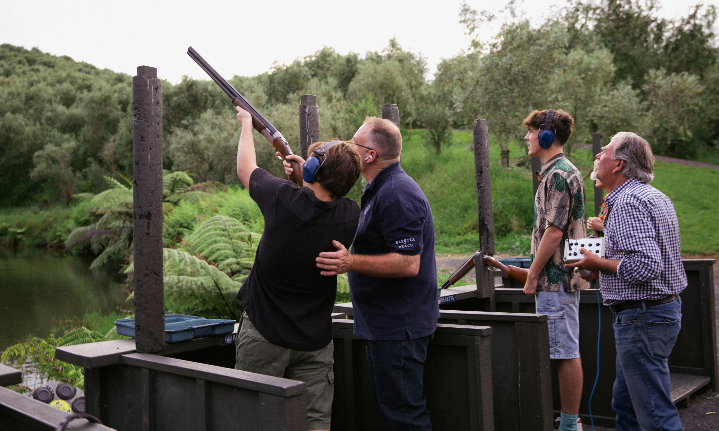 Four men at outdoor shooting range, with one aiming a shotgun, three observing, and one holding a remote control, surrounded by lush greenery and trees.