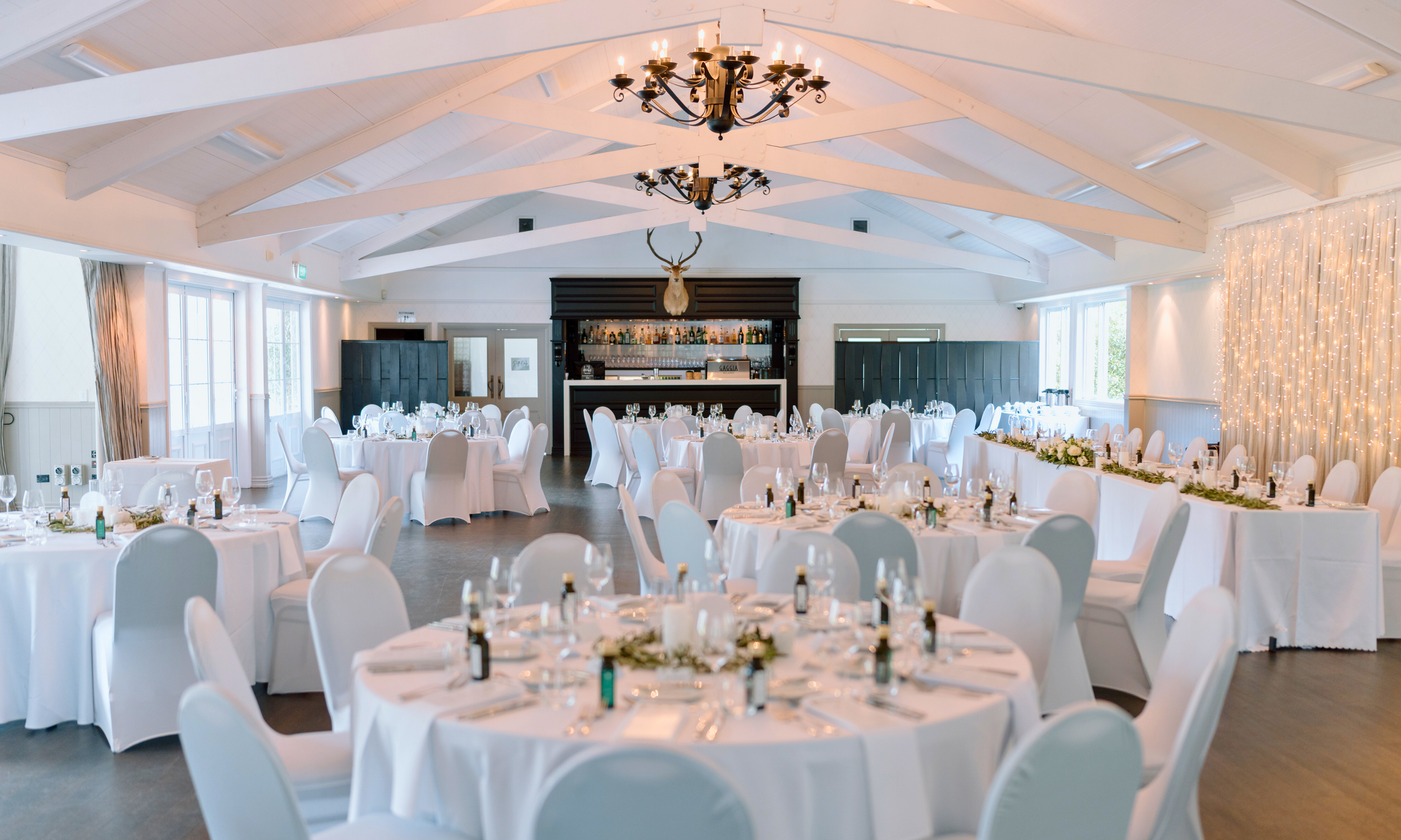 Elegant banquet hall decorated for a wedding or special event with white tablecloths, white chairs, and soft lighting, featuring a bar area with a mounted deer head and string lights on a wall.