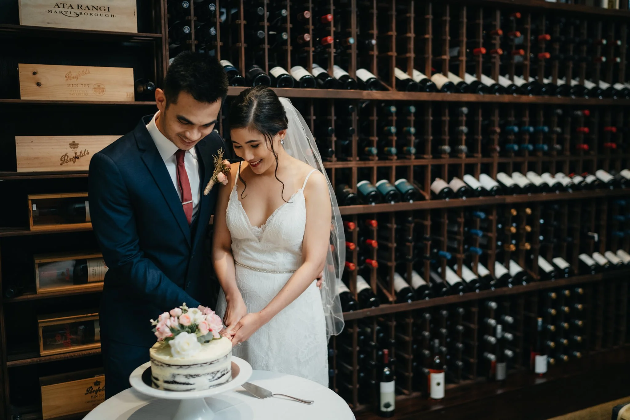 A newlywed couple, dressed in wedding attire, stands together cutting a wedding cake in a wine cellar.