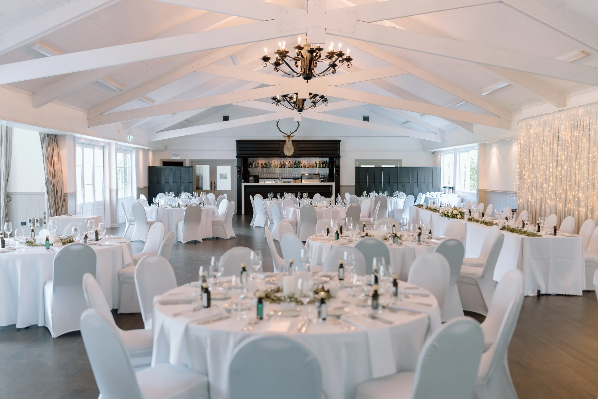 Elegant event hall decorated with round tables covered in white tablecloths, set for a formal dinner with white chairs, silverware, and glasses. The space features a high, white ceiling with exposed beams, large windows letting in natural light, and a backdrop with twinkling string lights. A chandelier hangs from the ceiling, and a mounted deer head is displayed above a bar area at the back of the room.