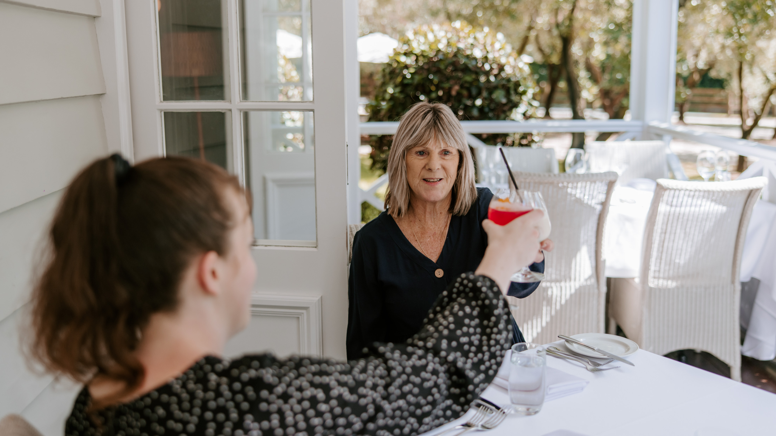 A woman is being offered a red cocktail with a straw by another woman at an outdoor dining area.