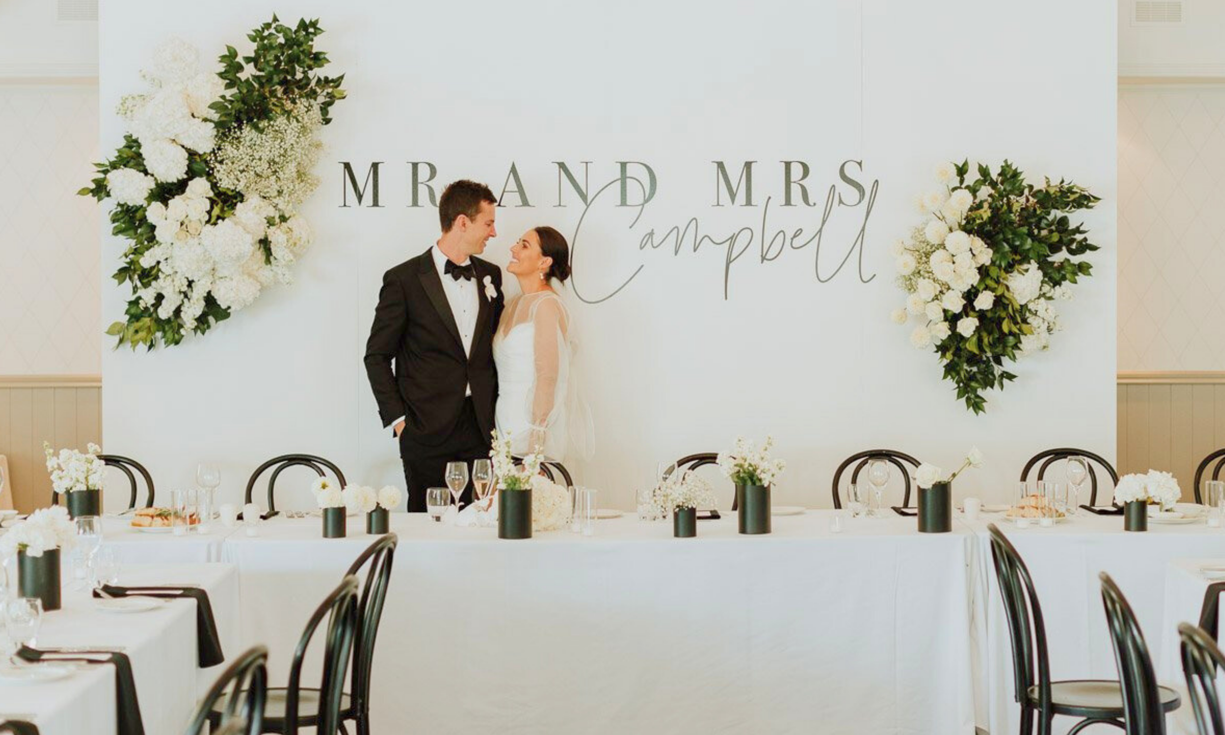 A bride and groom in wedding attire standing close together at a wedding reception, looking into each other's eyes, in front of a white backdrop with floral arrangements and the words 'Mr. and Mrs. Campbell' written on it.