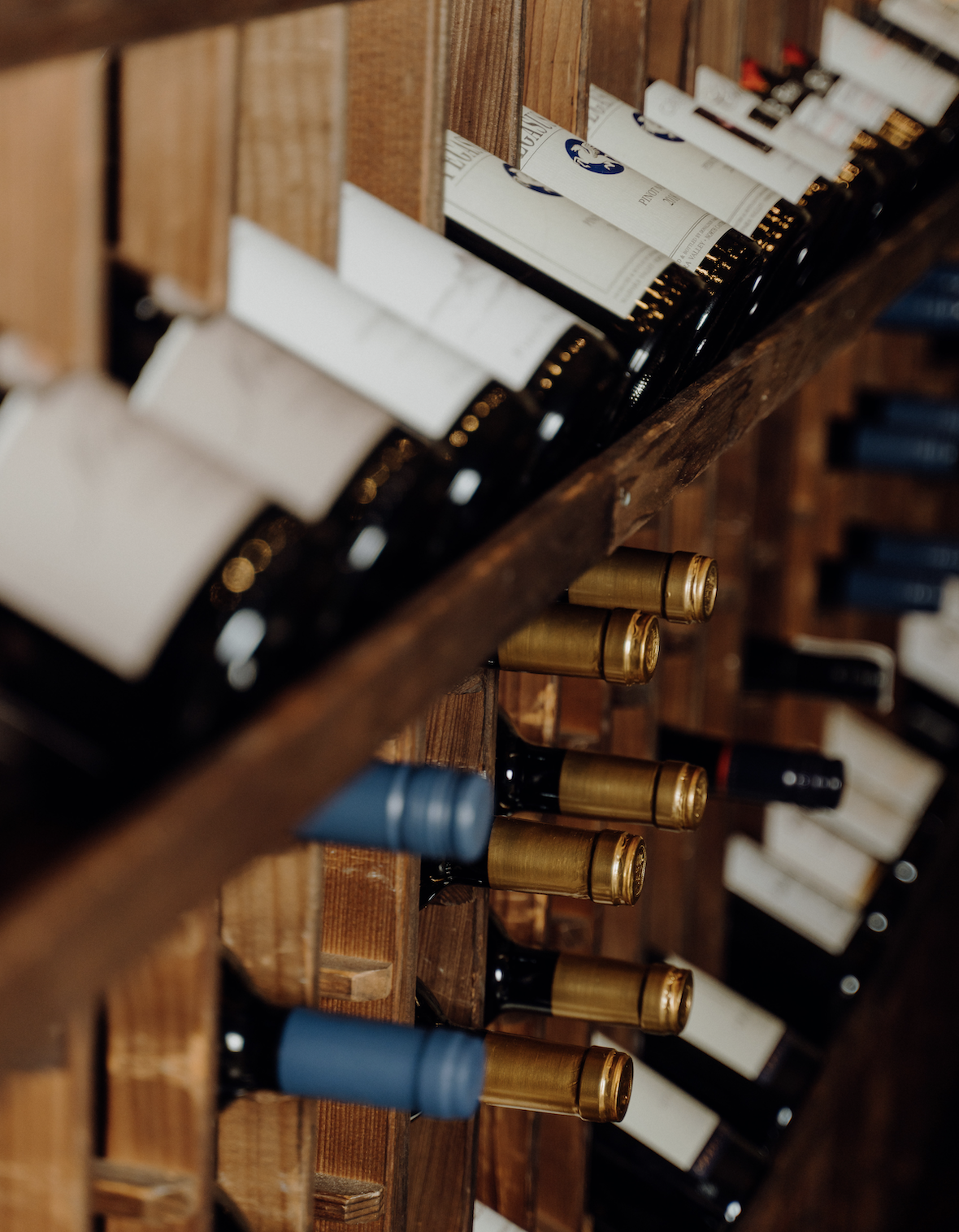 A wooden wine rack holding bottles of wine with gold and blue foil caps, displayed horizontally with labels facing outward.