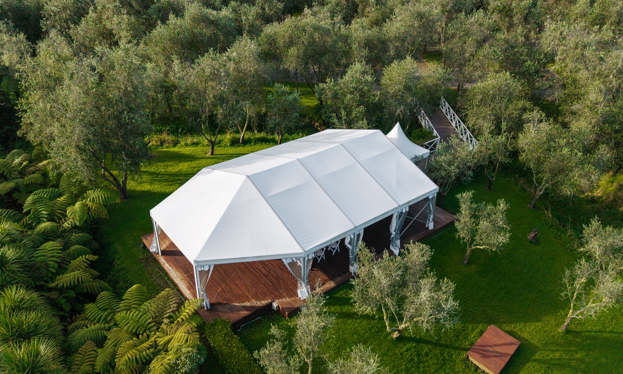 An aerial view of a large white tent with an elevated wooden deck surrounded by lush green trees and grass.