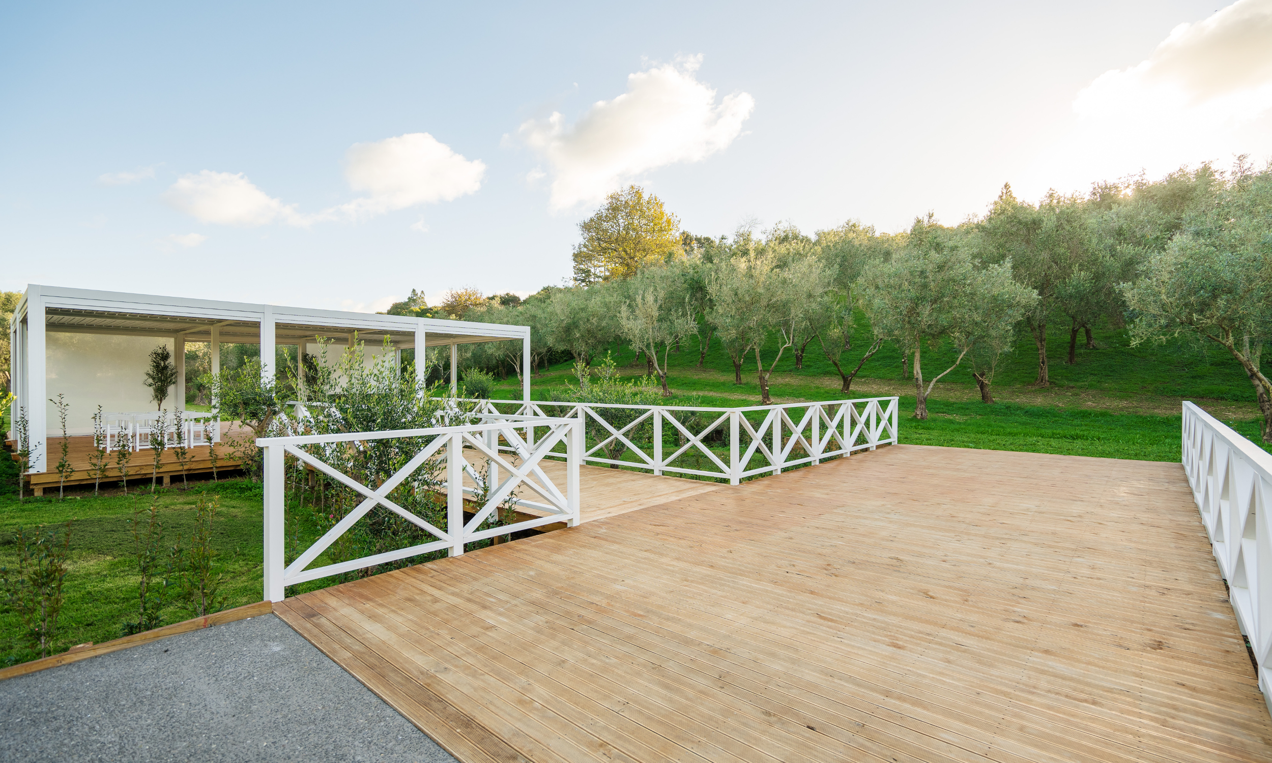 A wooden deck in a backyard with white railings, surrounded by green grass and trees, with a white outdoor structure and seating area in the background under a partly cloudy sky in the late afternoon.