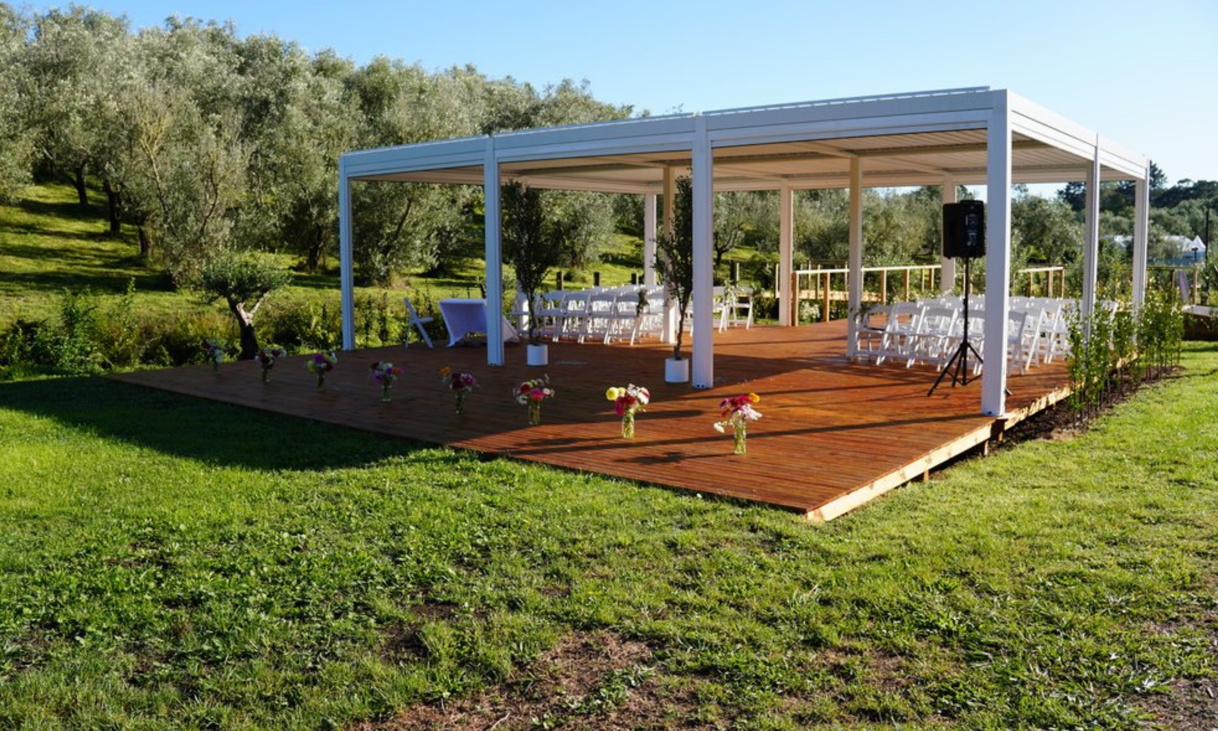 Outdoor wedding ceremony setup on a wooden platform with white chairs, decorated with flowers, under a white canopy, in a lush green landscape with trees and grass.