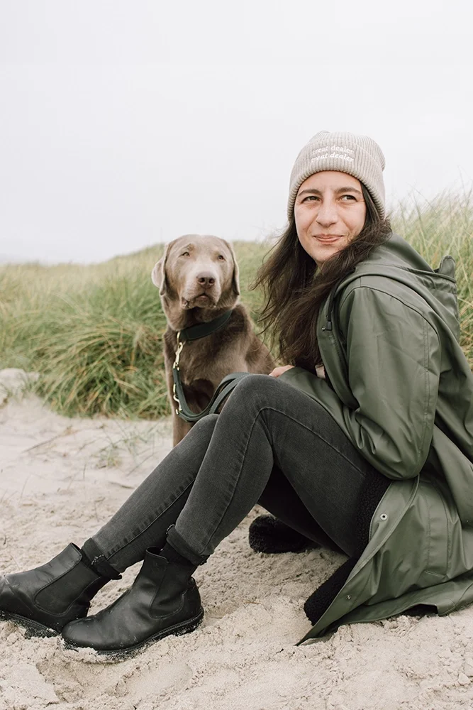 Eine Frau sitzt mit einem braunen Hund an einem Strand, umgeben von Sand und Gras, bei bewölktem Himmel.