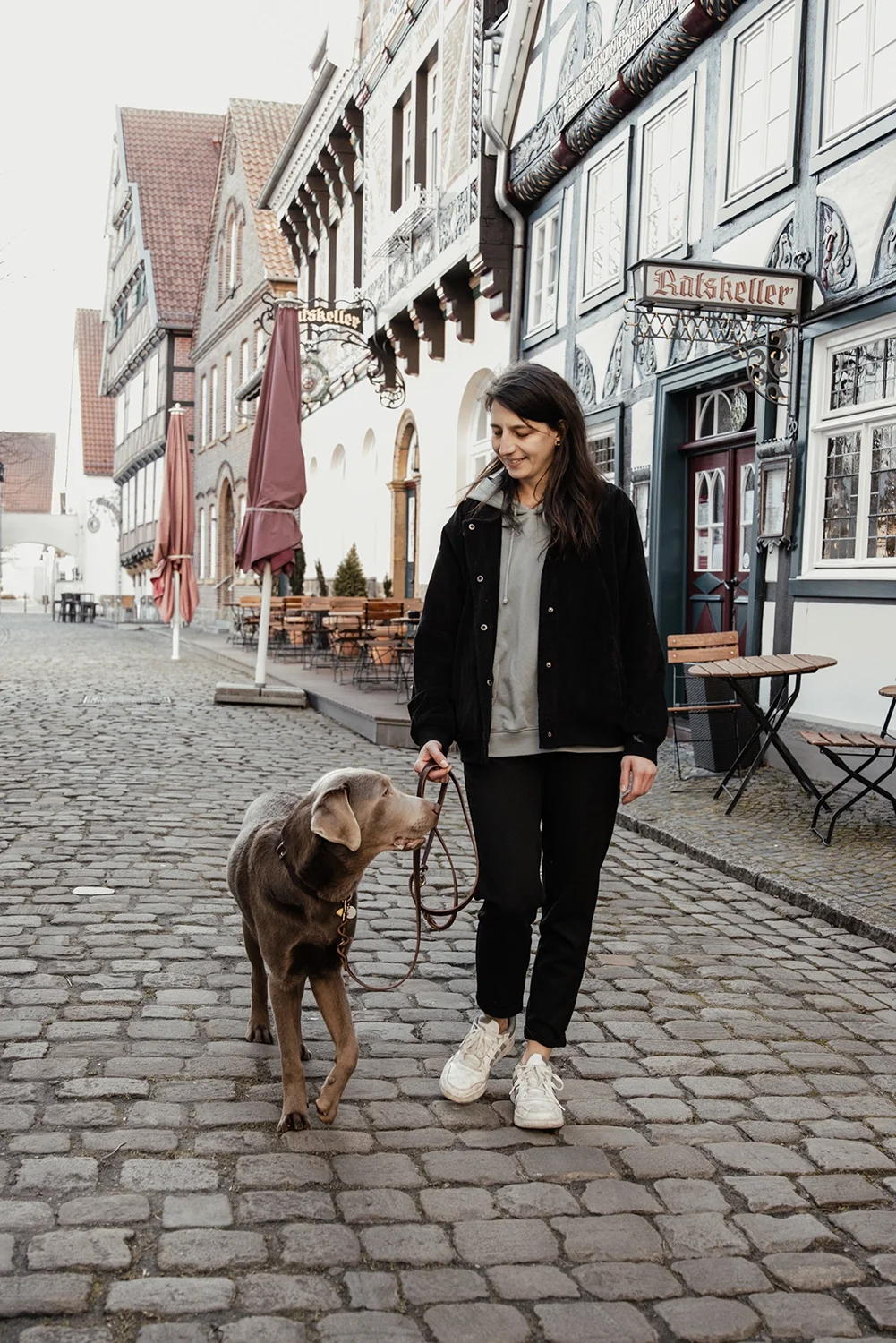 Eine junge Frau geht mit einem braunen Hund auf einem Kopfsteinpflaster in einer historischen europäischen Stadt. Im Hintergrund sind alte Gebäude mit Fassaden im Fachwerkstil und eine Gaststättentafel mit der Aufschrift 'Ratskeller' zu sehen.