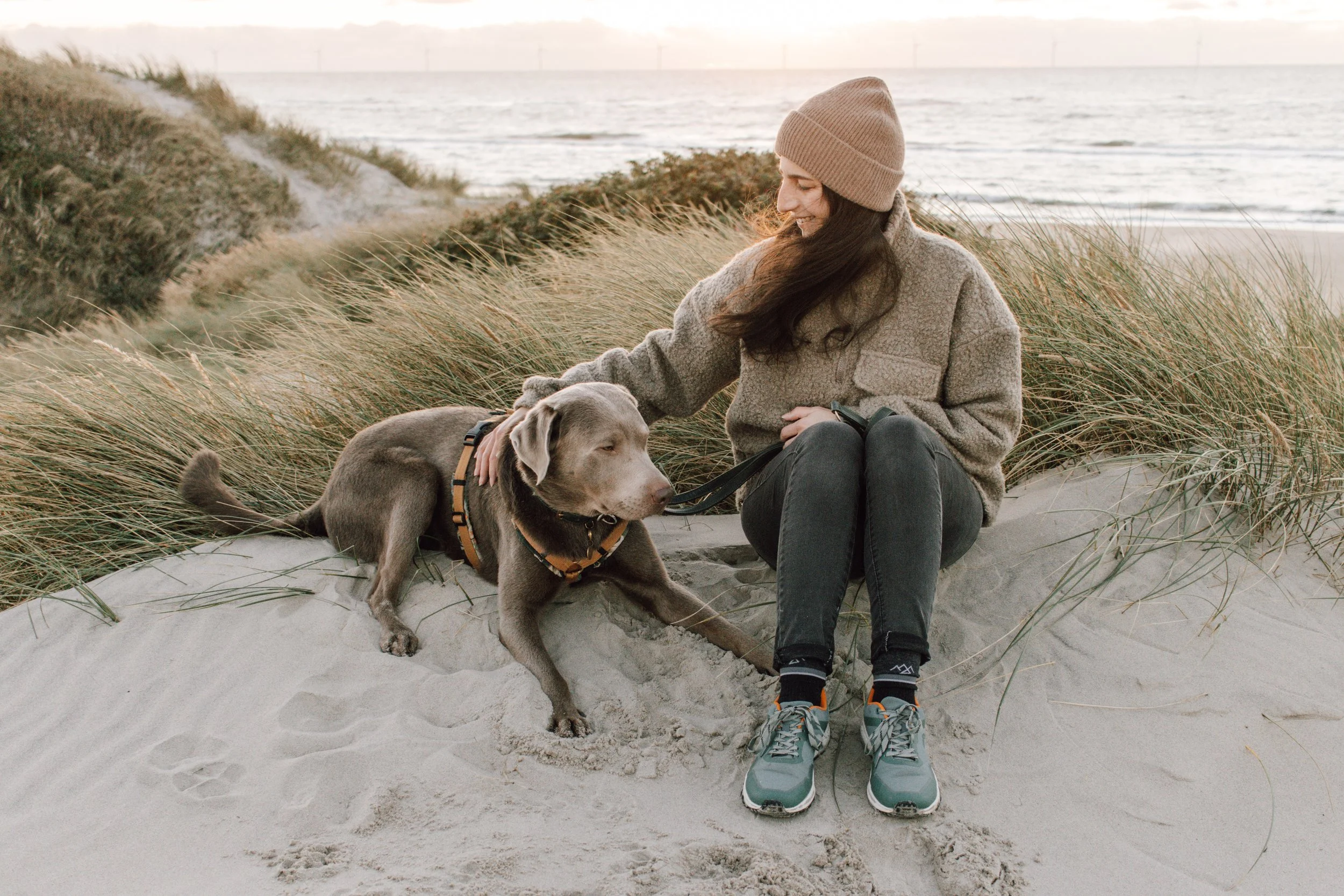 Eine junge Frau mit braunem Hut und grauem Pullover sitzt am Strand auf Sand und umgibt sich mit Dünen und Gräsern. Sie streichelt einen grauen Hund, der neben ihr liegt, während im Hintergrund das Meer und ein Sonnenuntergang zu sehen sind.