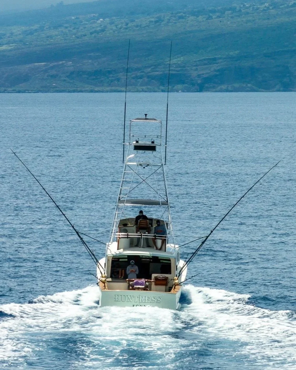 A boat named 'HUNTRESS' on the water with people fishing, in a scenic location with distant mountains.