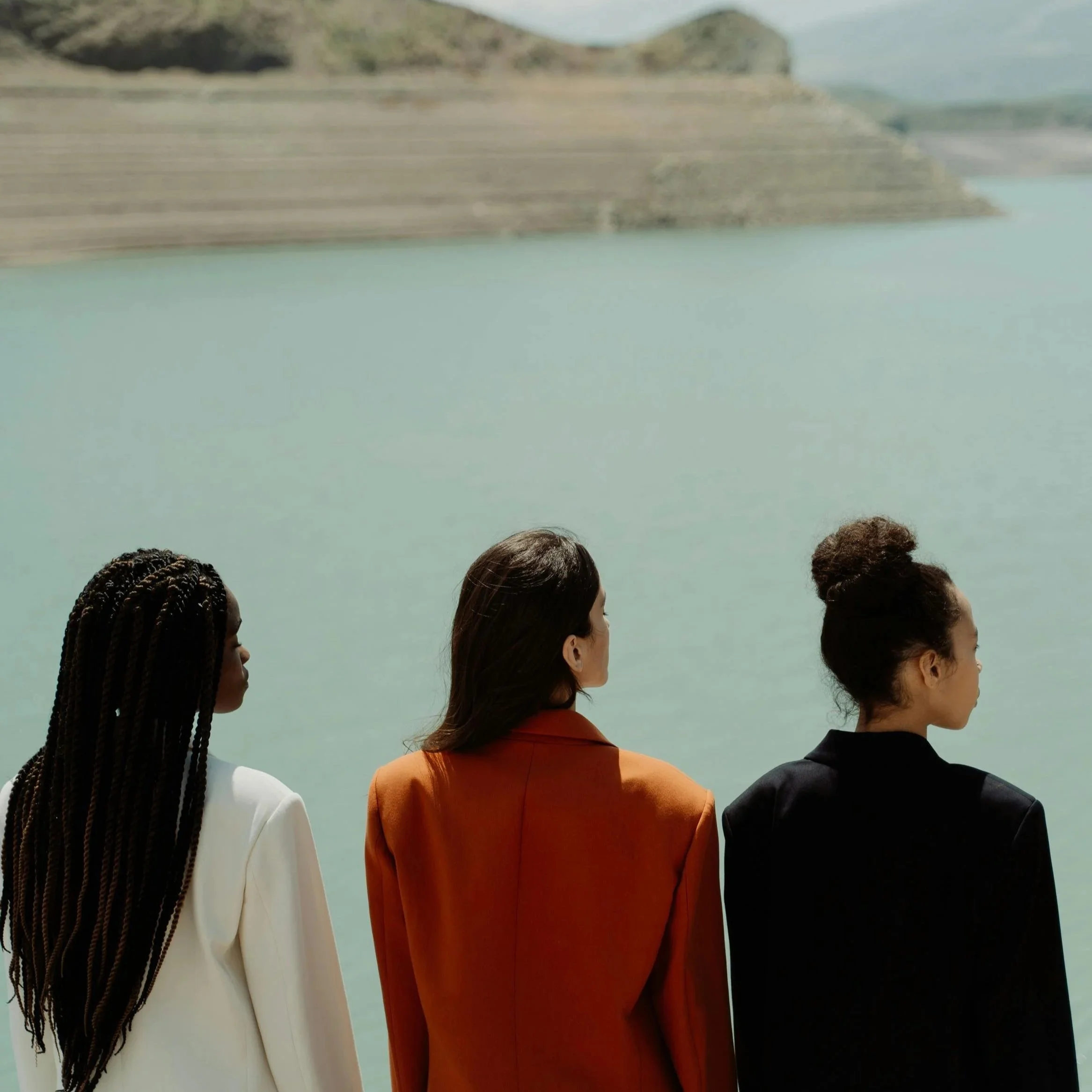Three women standing by the water, facing away, with a landscape of a lake and hills in the background.