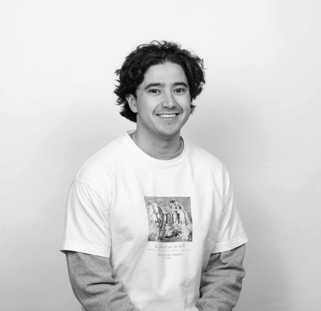 Black and white portrait of a young man with dark, curly hair smiling, wearing a white graphic t-shirt layered over a long-sleeve shirt against a plain background.