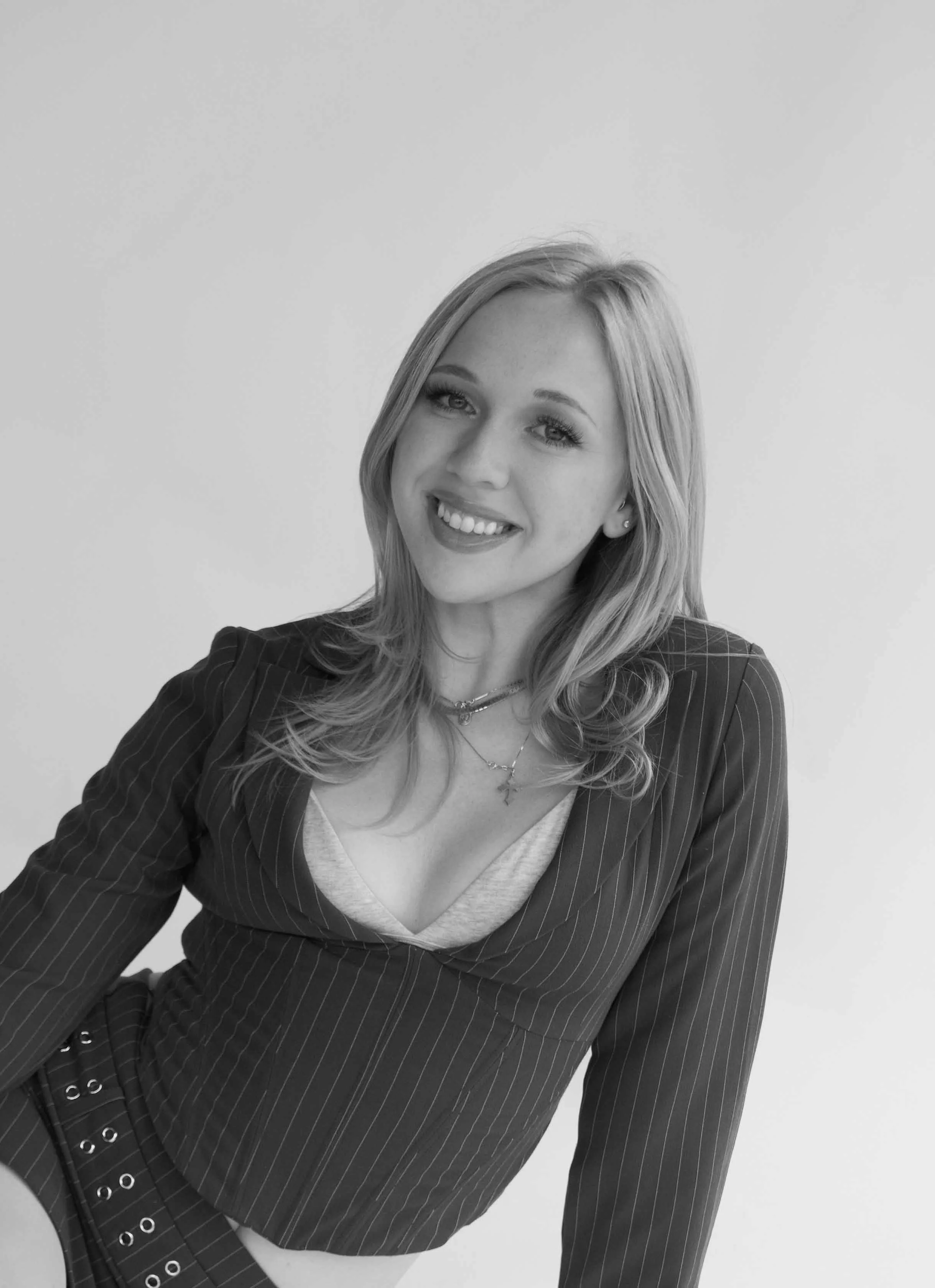 Black and white photo of a young woman with long wavy hair, smiling, wearing a striped blazer over a top with a deep neckline, and layered necklaces, sitting against a plain background.