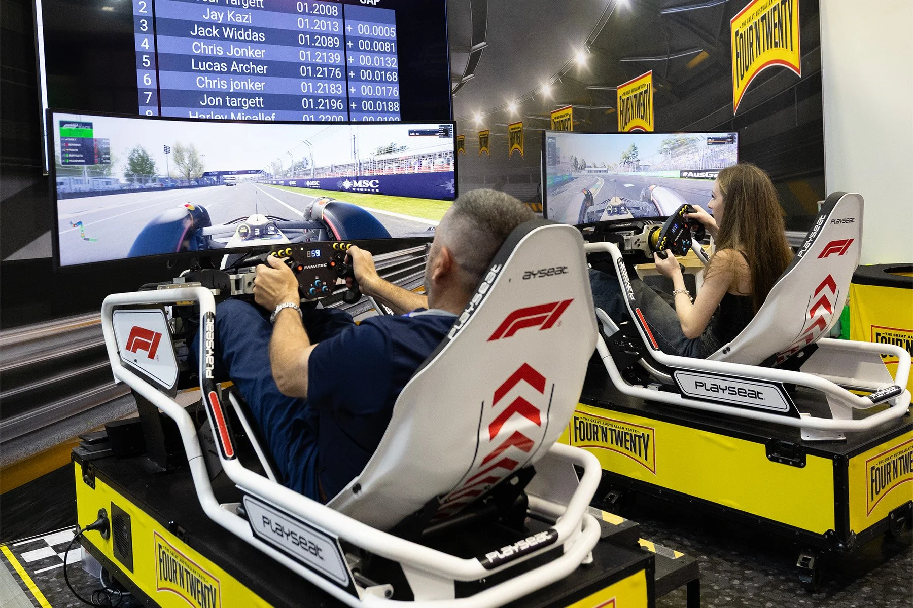 Two people are racing in F1 racing simulators, seated in yellow and black PlaySeat racing chairs with F1 logos, with large screens displaying a NASCAR race and a racing game, and a leaderboard showing race times and positions in the background.
