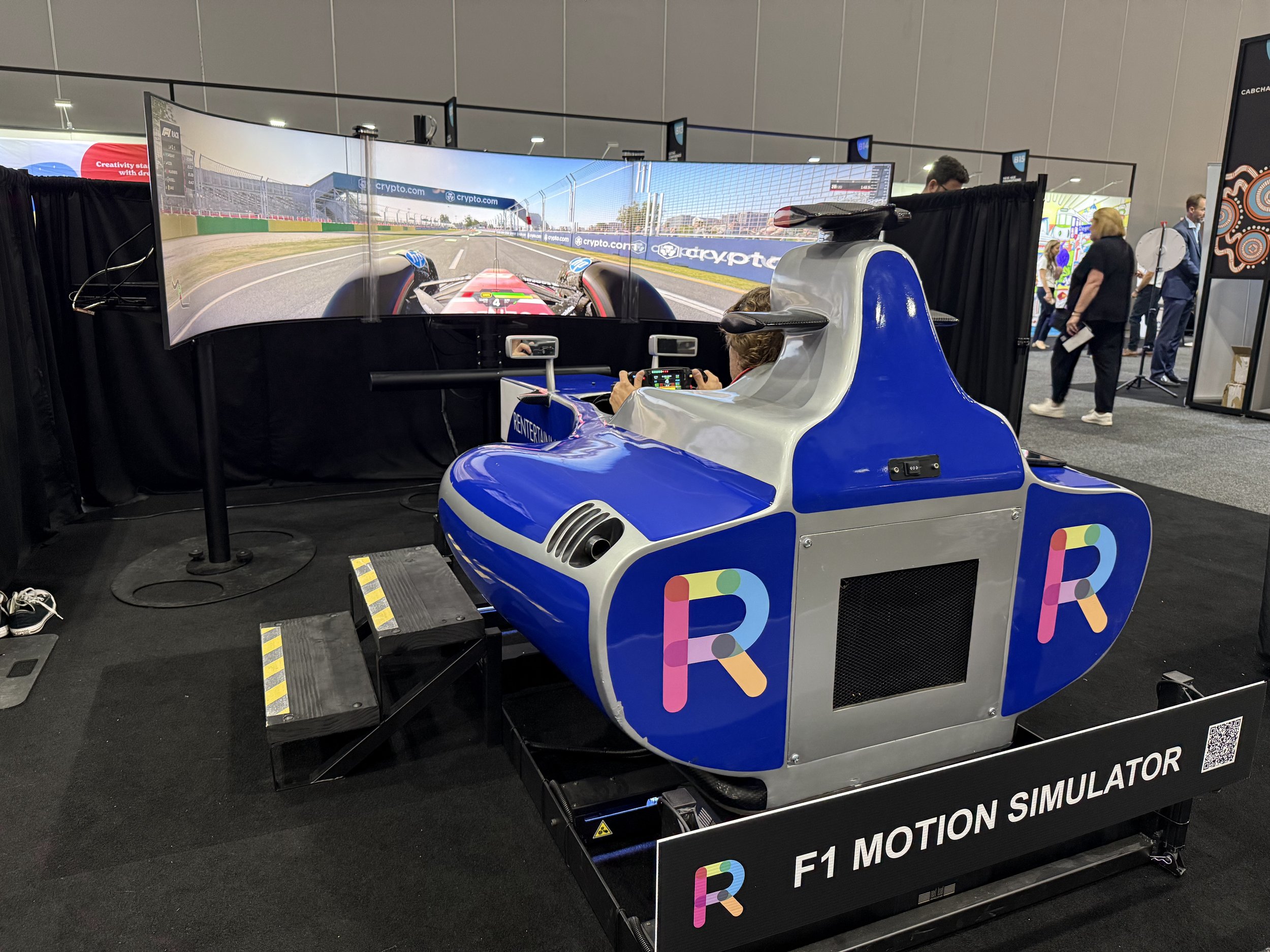 A person sitting in a blue and gray F1 racing car motion simulator at a technology expo, with a large curved screen displaying a racing game in front of them.