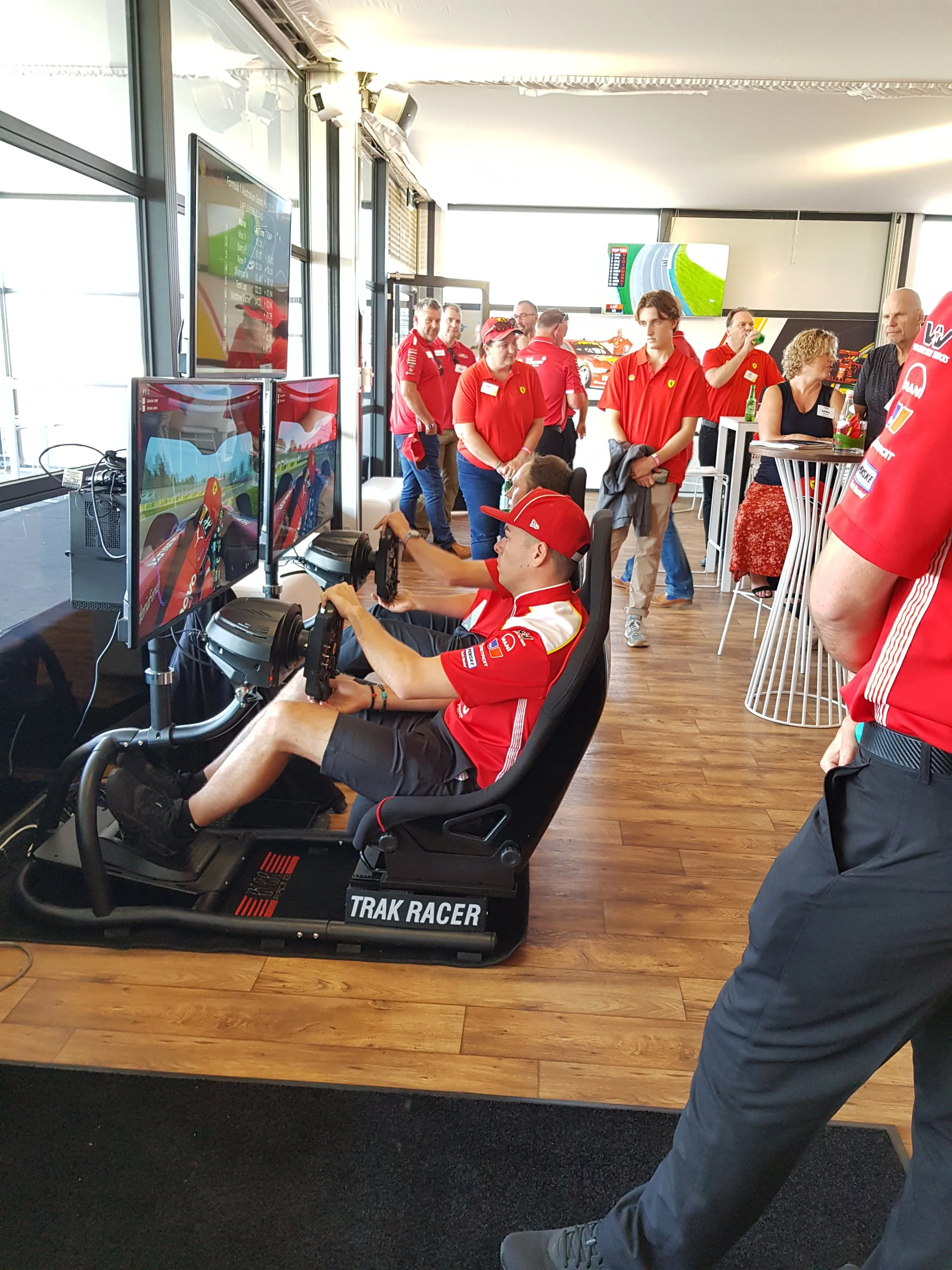 A young person wearing a red cap and racing shirt is sitting in a racing simulator labeled "TRAK RACER" with multiple monitors showing a racing game. Several people, mostly in red shirts, are standing and watching in the background, in a room with large windows and wooden flooring.