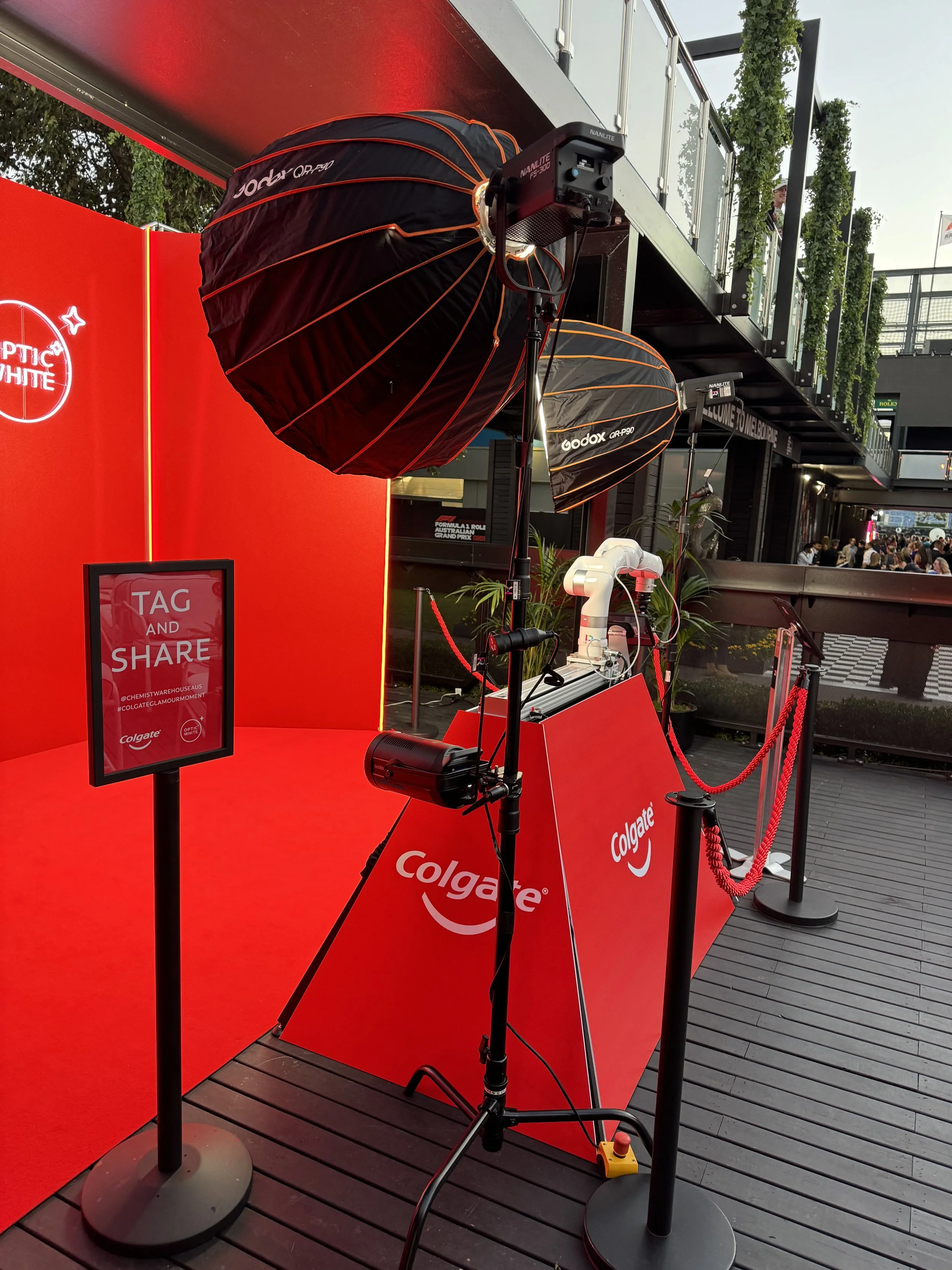 Photography setup with lighting equipment at a Colgate event, including large umbrella lights and a red backdrop with promotional signage reading 'TAG AND SHARE' and 'Colgate' logo.
