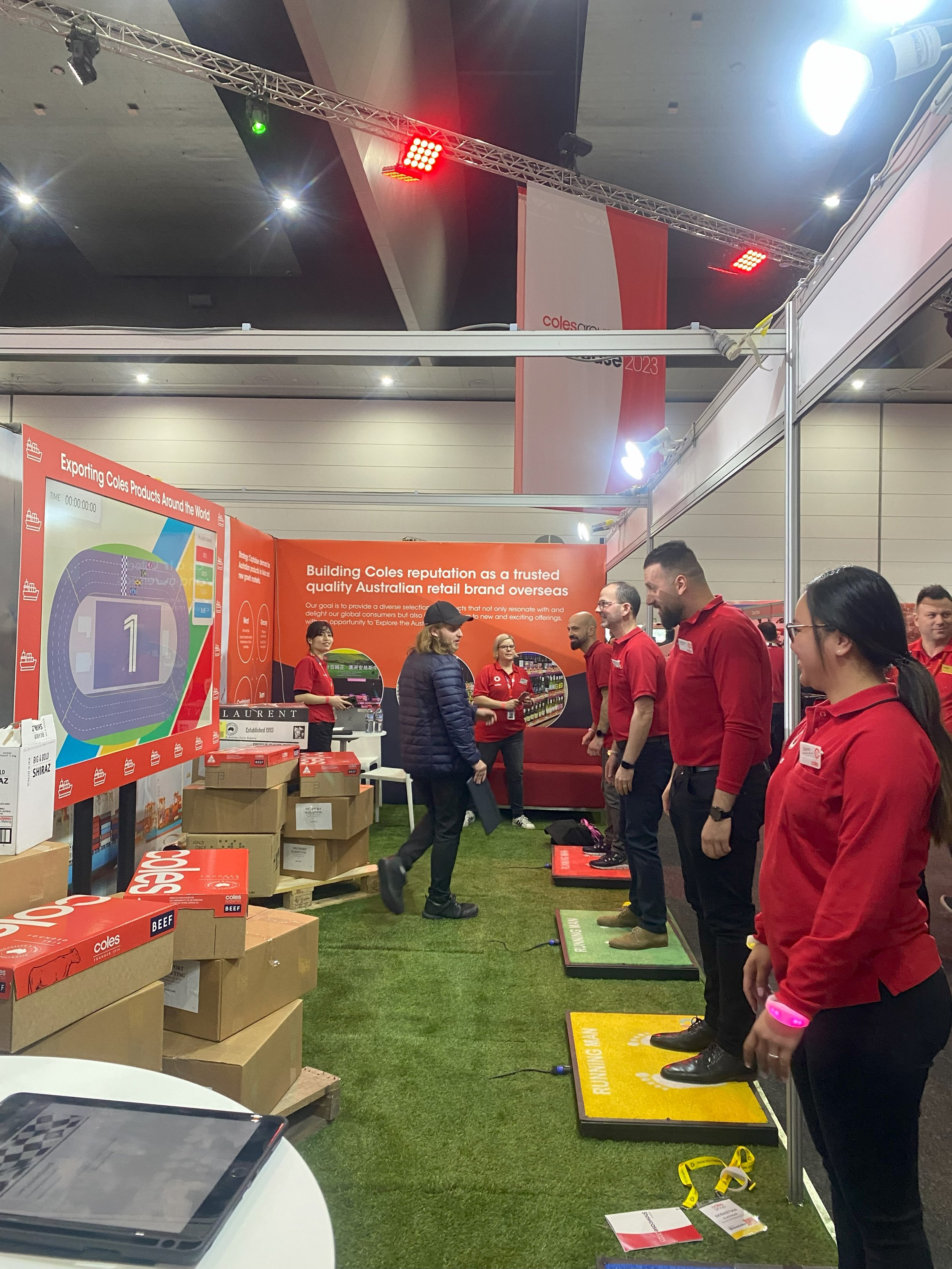 Group of people at a trade show booth, some standing on foot pedals, demonstrating a product or activity related to Coles, an Australian retail brand. The booth has red branding and promotional displays.