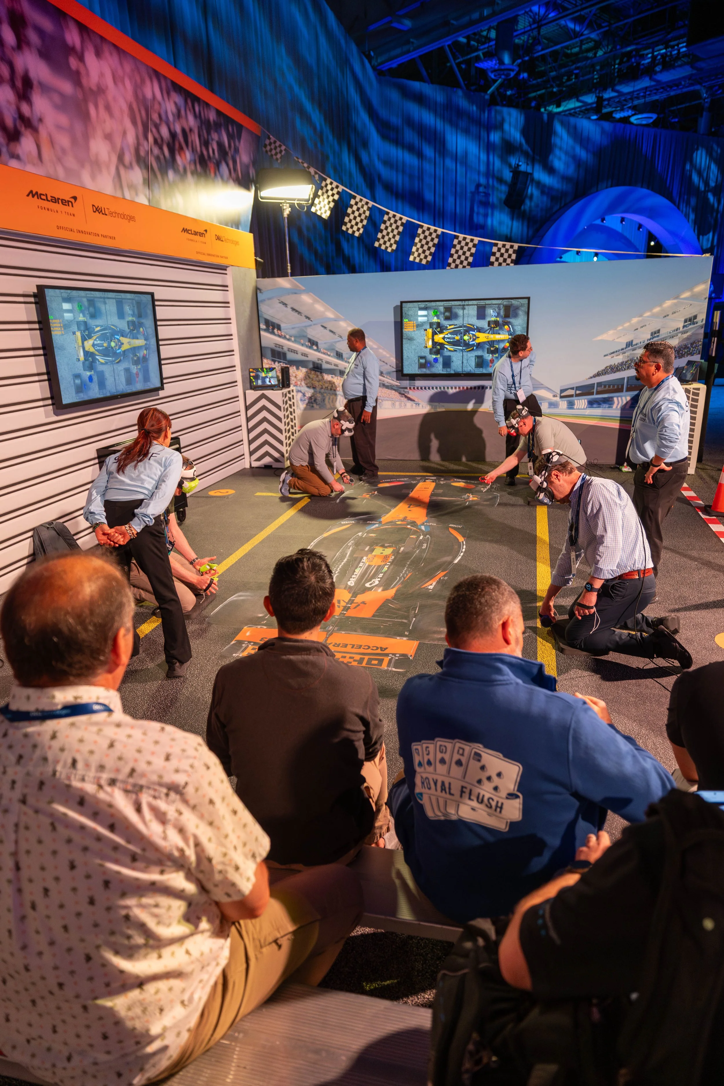 People gather around a race car simulation setup with virtual reality headsets at a motorsport event, with multiple screens displaying racing scenes and a large backdrop of a racetrack.
