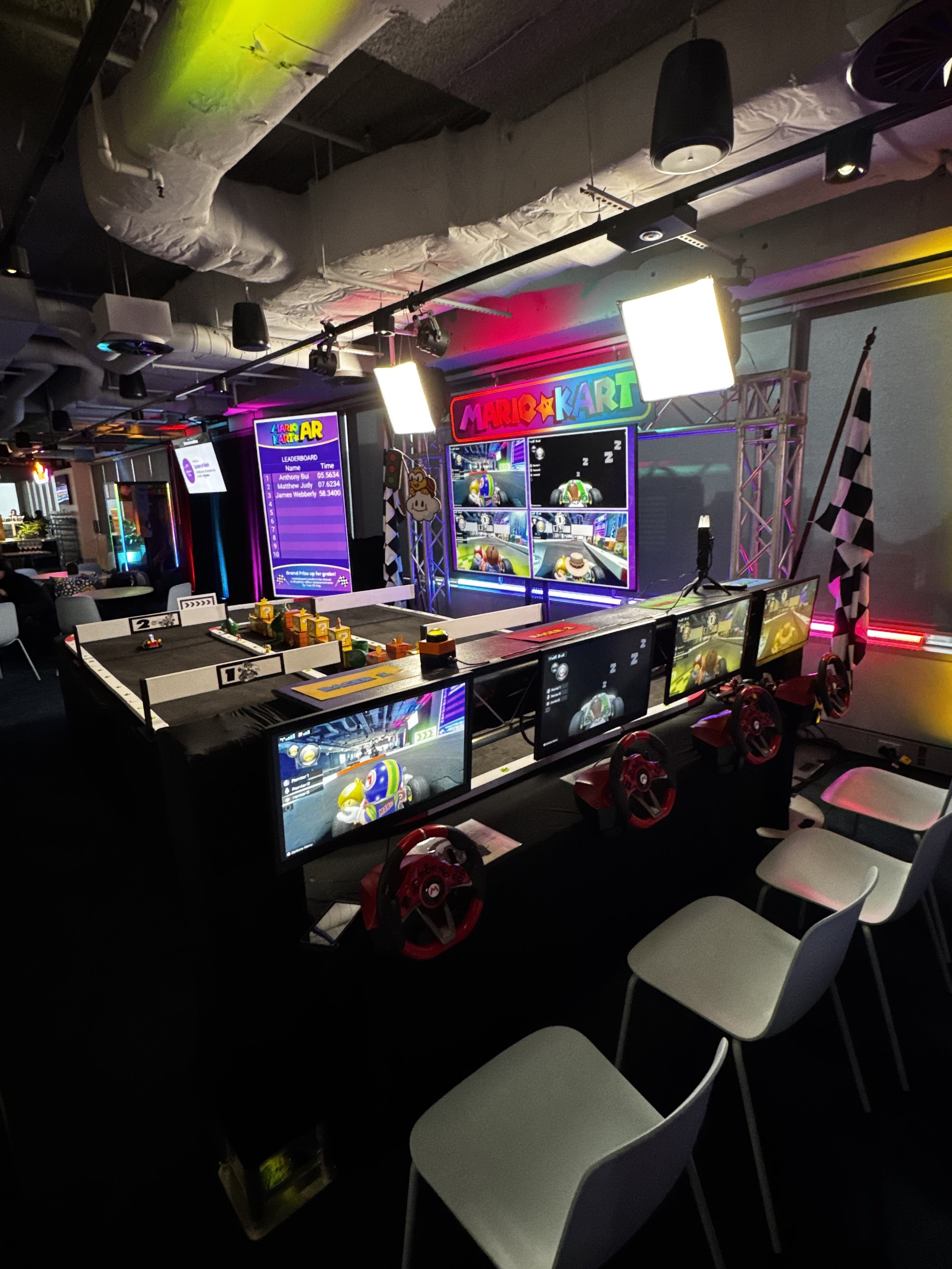 Indoor arcade gaming area with Mario Kart racing setup, multiple monitors, racing wheels, and television screens displaying gameplay, with chairs in front and leaderboard on a digital screen.