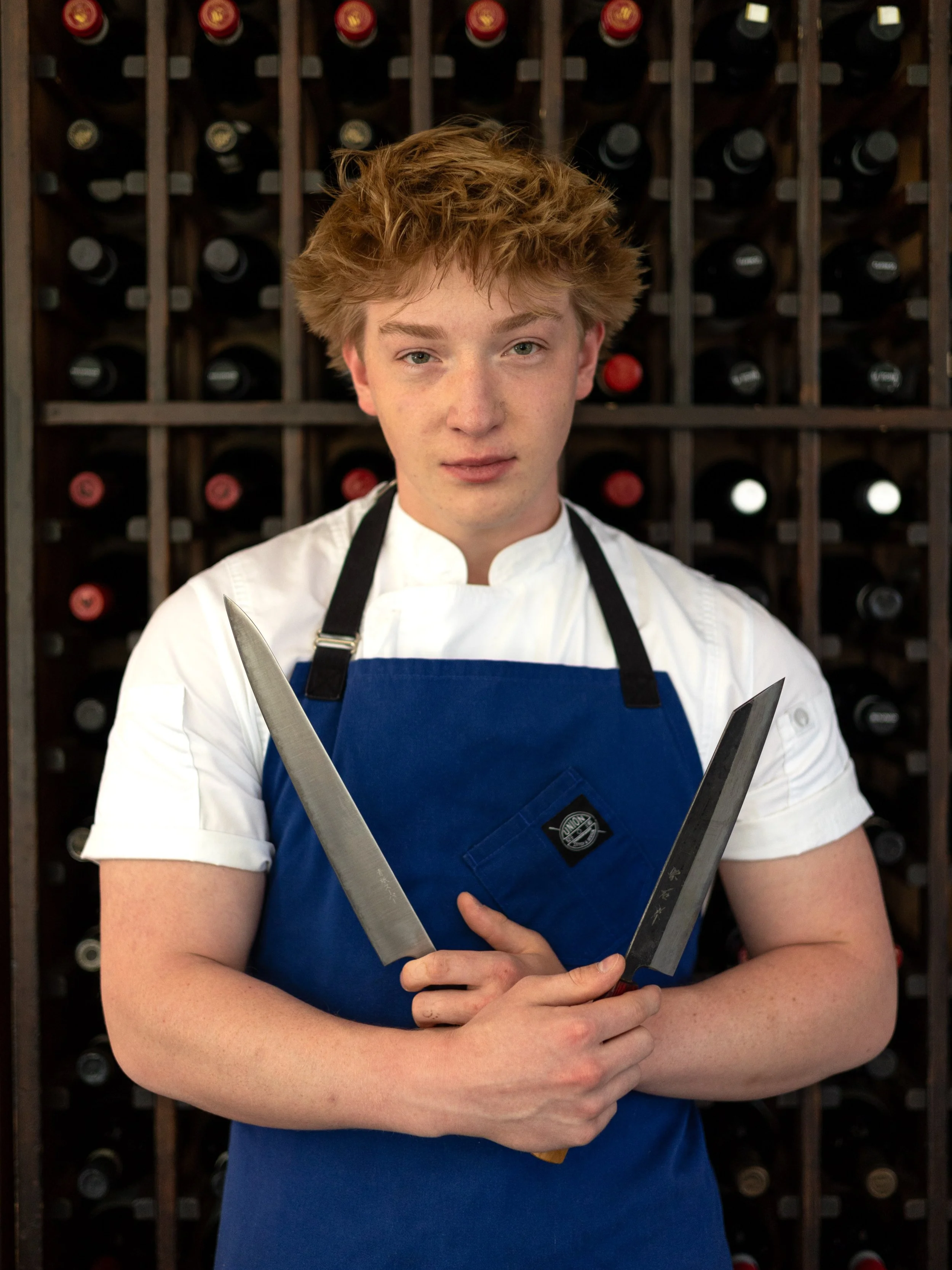 A young man with short, blonde hair, wearing a white chef's coat and a blue apron, holds two large kitchen knives crossed in front of him. He stands in front of a wine rack full of bottles.