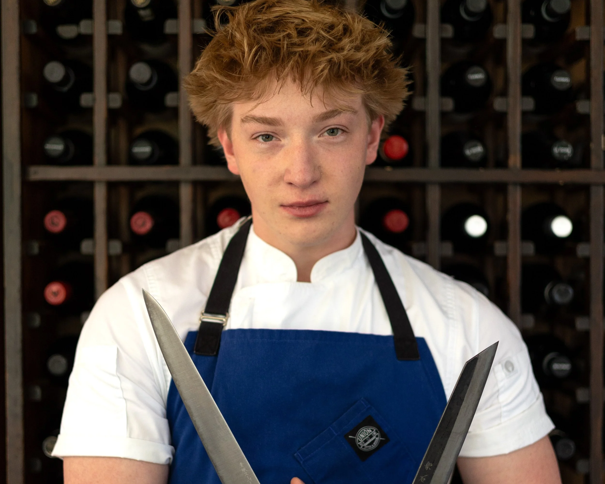 A young man with short, blonde hair, wearing a white chef's coat and a blue apron, holds two large kitchen knives crossed in front of him. He stands in front of a wine rack full of bottles.