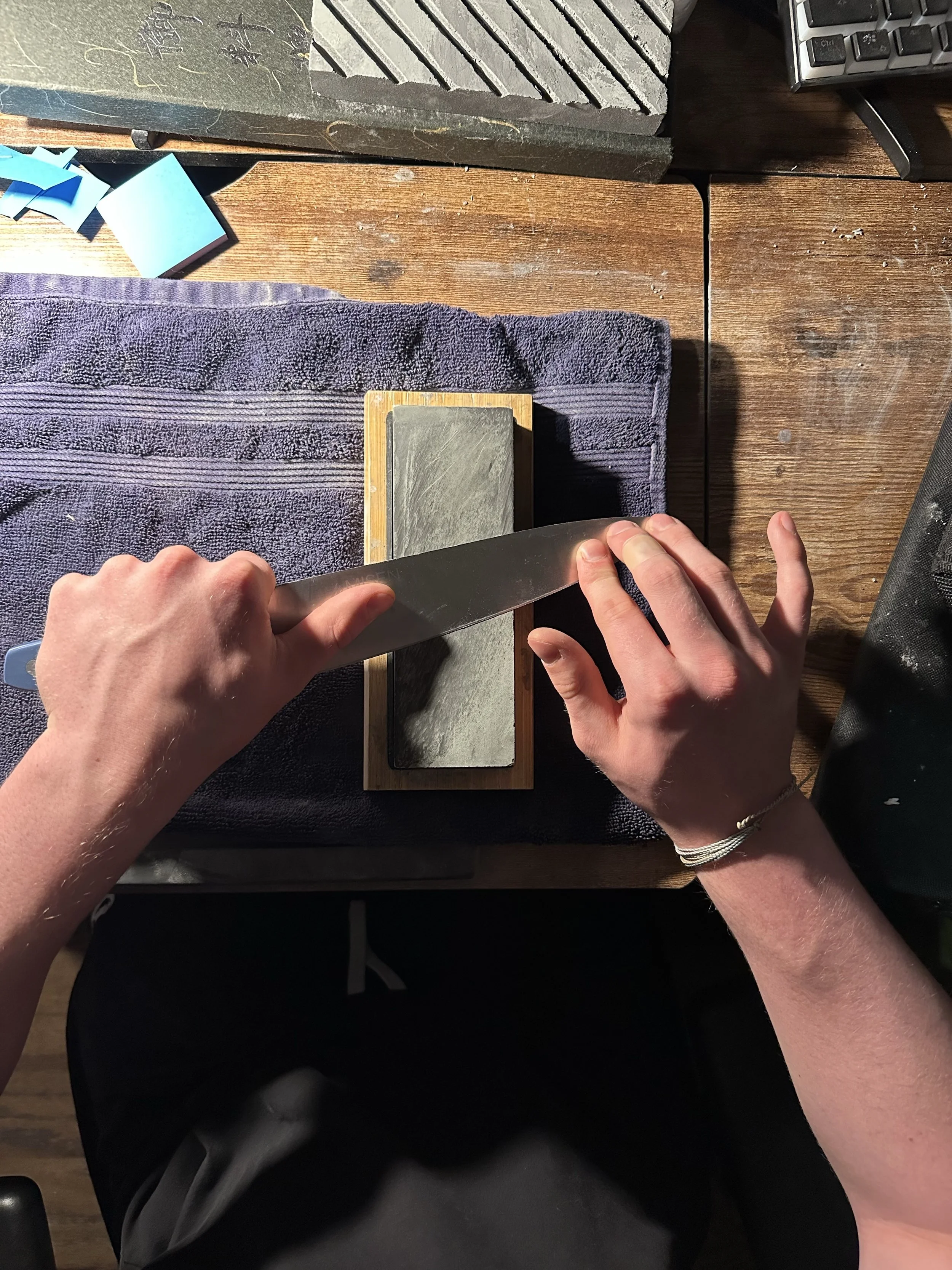 Person sharpening a knife on a sharpening stone with a black sharpening guide, wooden base, and a blue towel on a wooden desk.